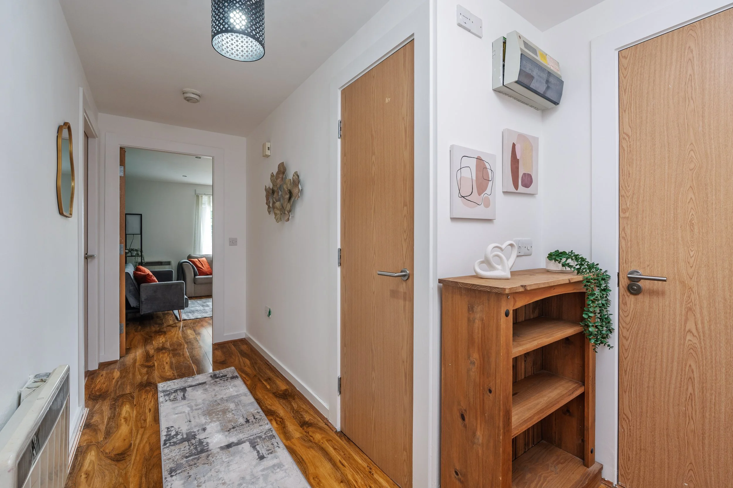 Hallway leading to a living room with two armchairs and a window. The hallway has white walls, wood flooring, a gray patterned rug, and decorative wall art. On the right, there's a wooden shelf with plants and abstract wall art, and doors with wood finish. A black ceiling light fixture is visible.