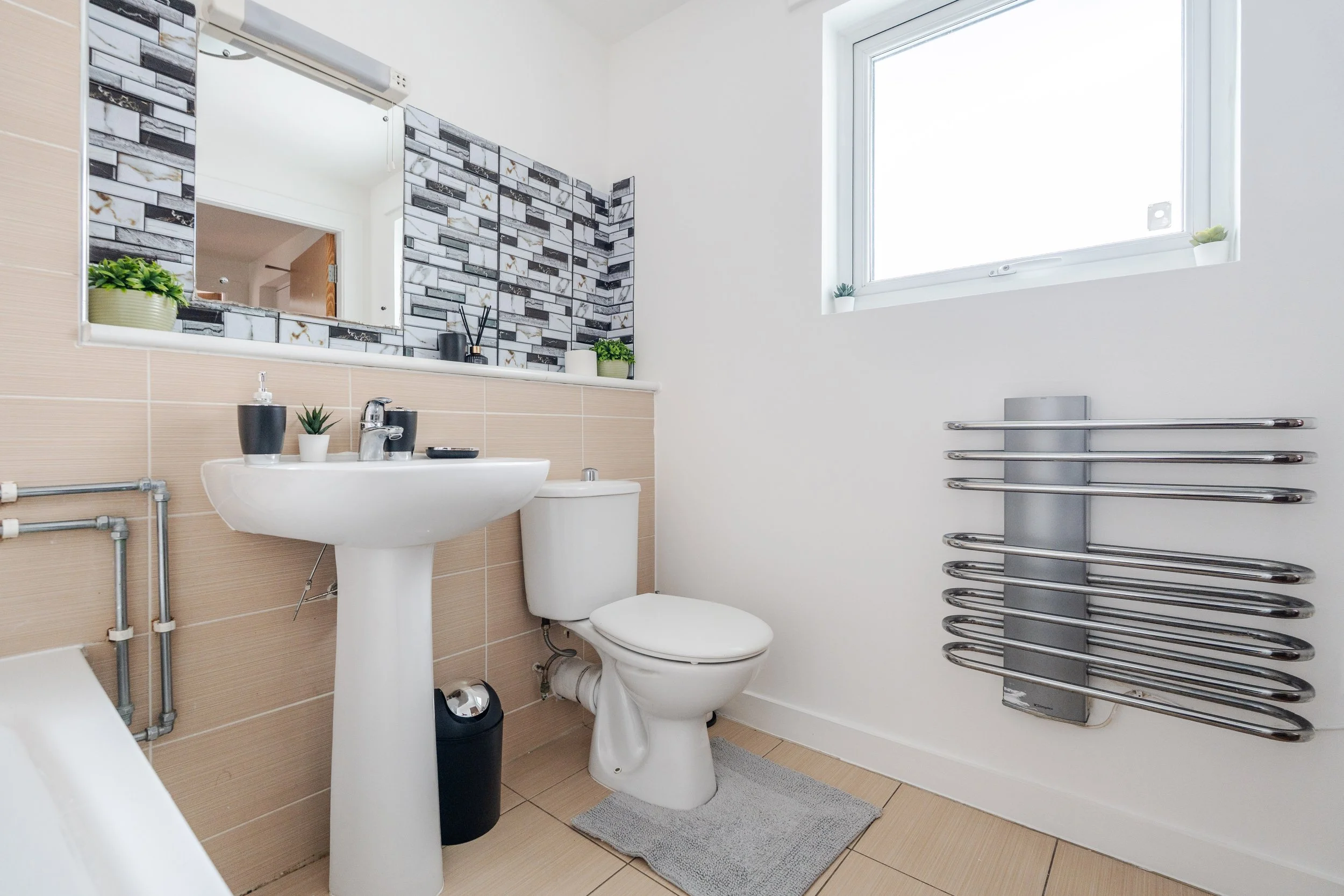 A modern bathroom with beige tiled walls and a white toilet. There is a white pedestal sink with a small plant and black soap dispenser. A large mirror above the sink reflects a decorated wall with black, white, and gray tile design. To the right is a large window with small potted plants on the sill. A metal towel radiator is mounted on the right wall. The bathroom has a light gray rug on the beige tiled floor.