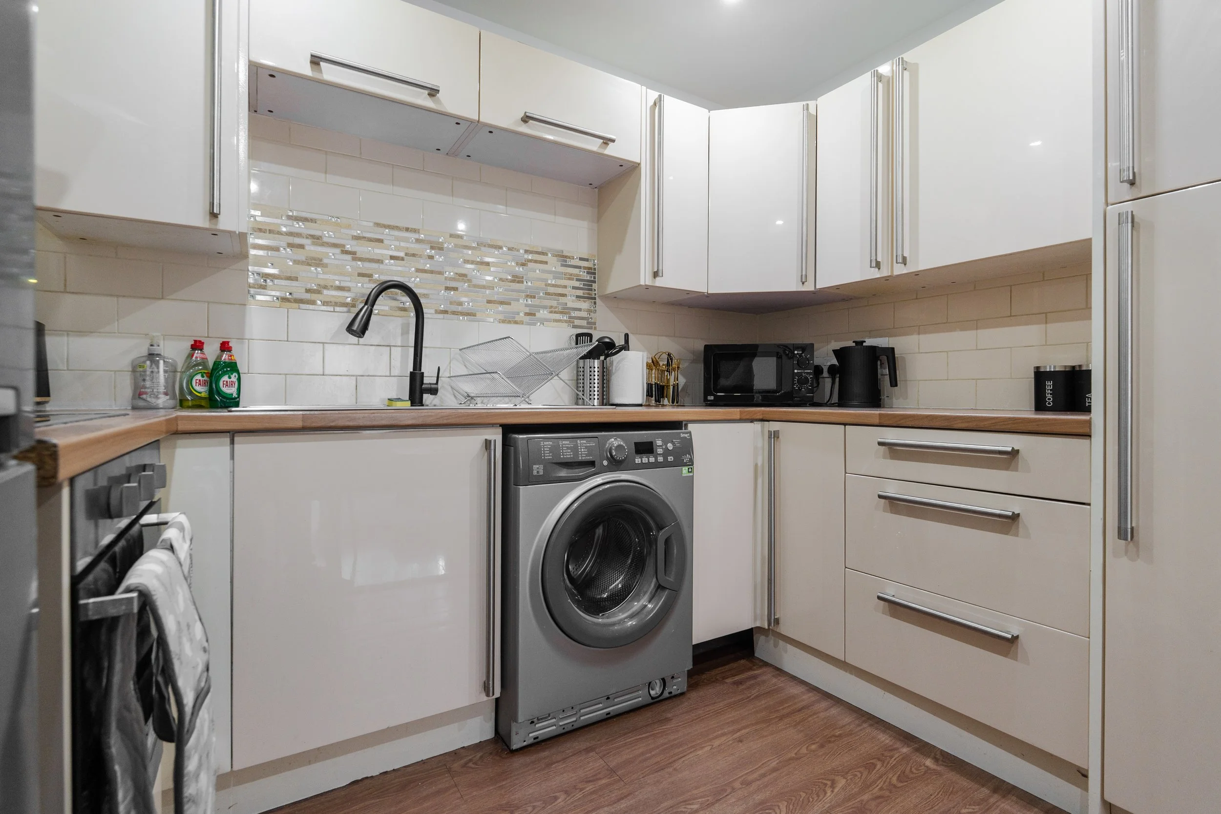 A compact kitchen with white cabinets, a wooden countertop, a gray washing machine, a black microwave, and various small appliances and cleaning supplies on the counter.