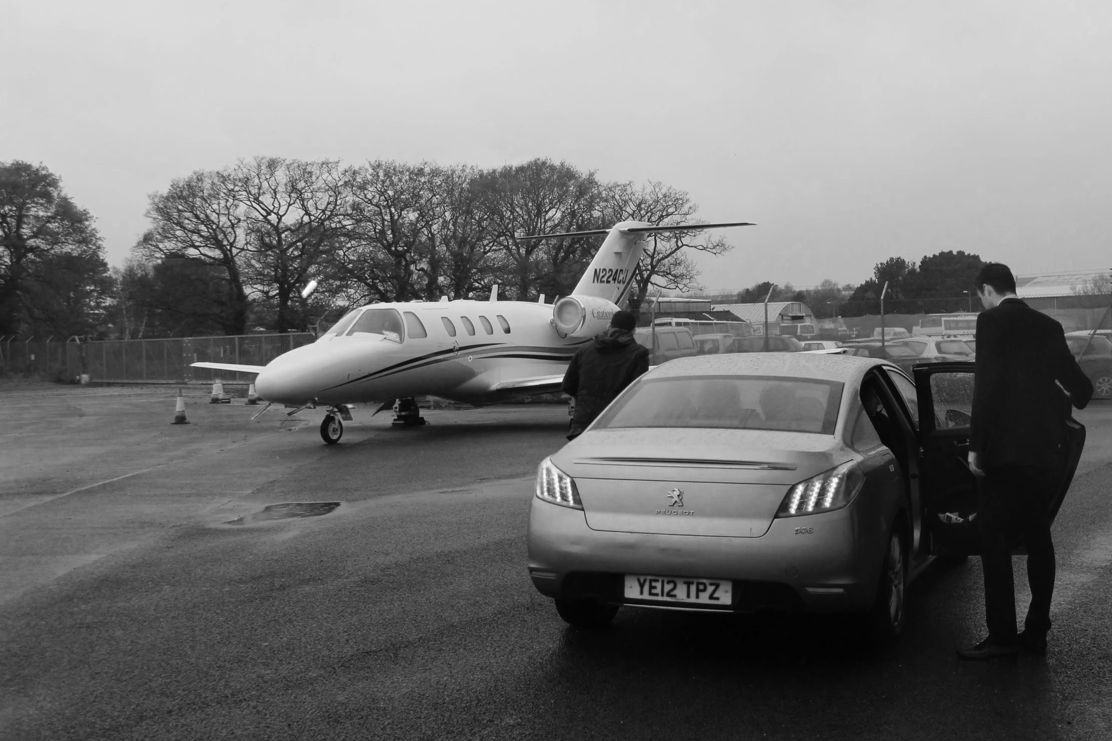 Black and white photo of a small private jet parked on a tarmac with two men nearby, one opening a car door and the other standing next to a parked car. Trees and a fence are visible in the background.
