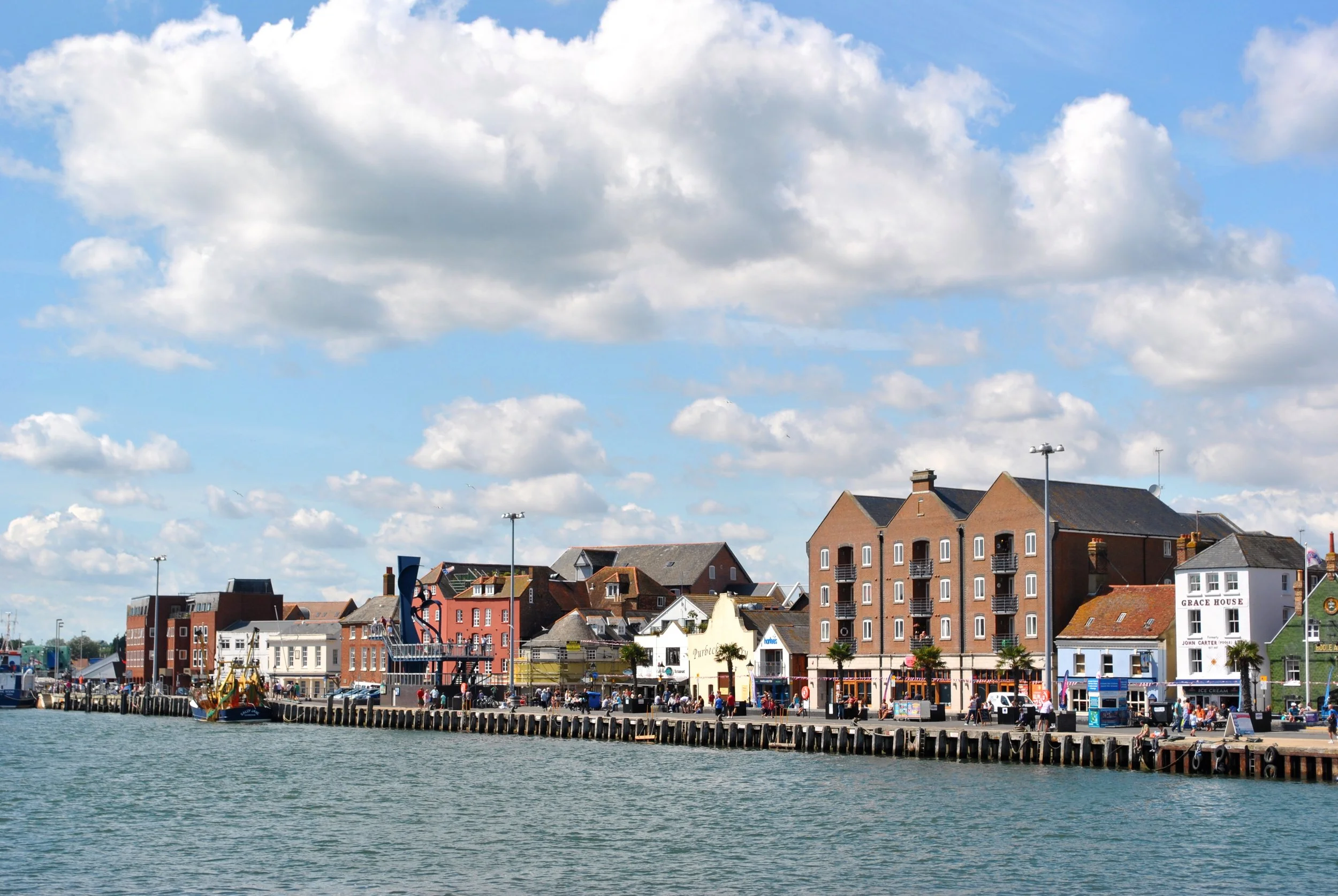 A waterfront scene with a pier in the foreground, colorful buildings including residential and commercial establishments along the shoreline, and a partly cloudy sky overhead.