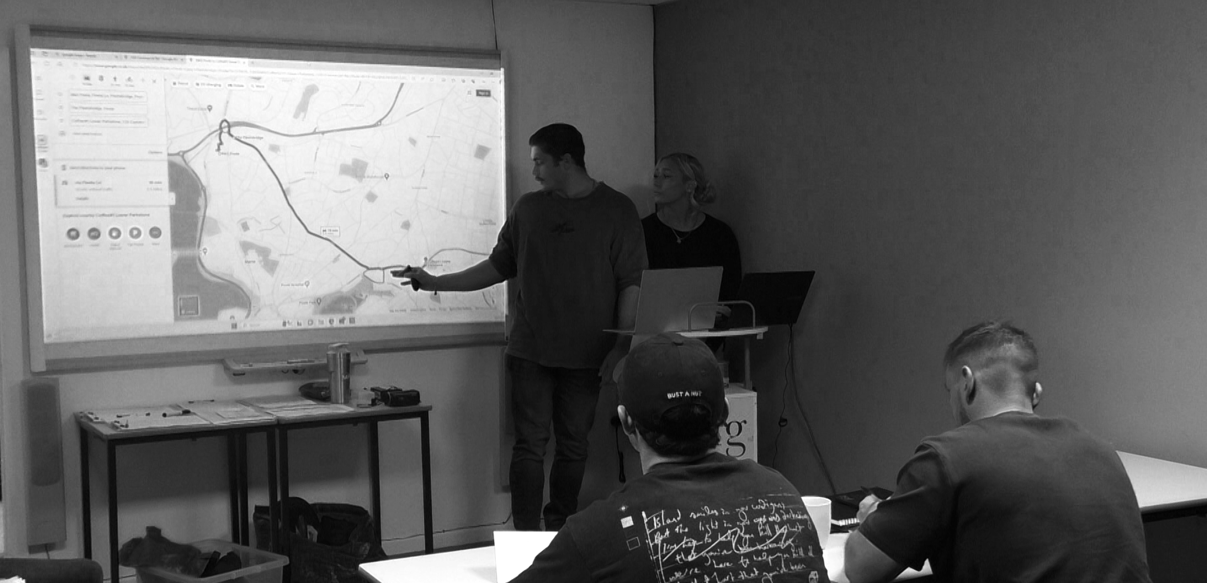 Two people are giving a presentation in a room with a projected map on a whiteboard. The man on the left is pointing at the map, while the woman on the right is standing nearby. Several attendees are seated, listening to the presentation.