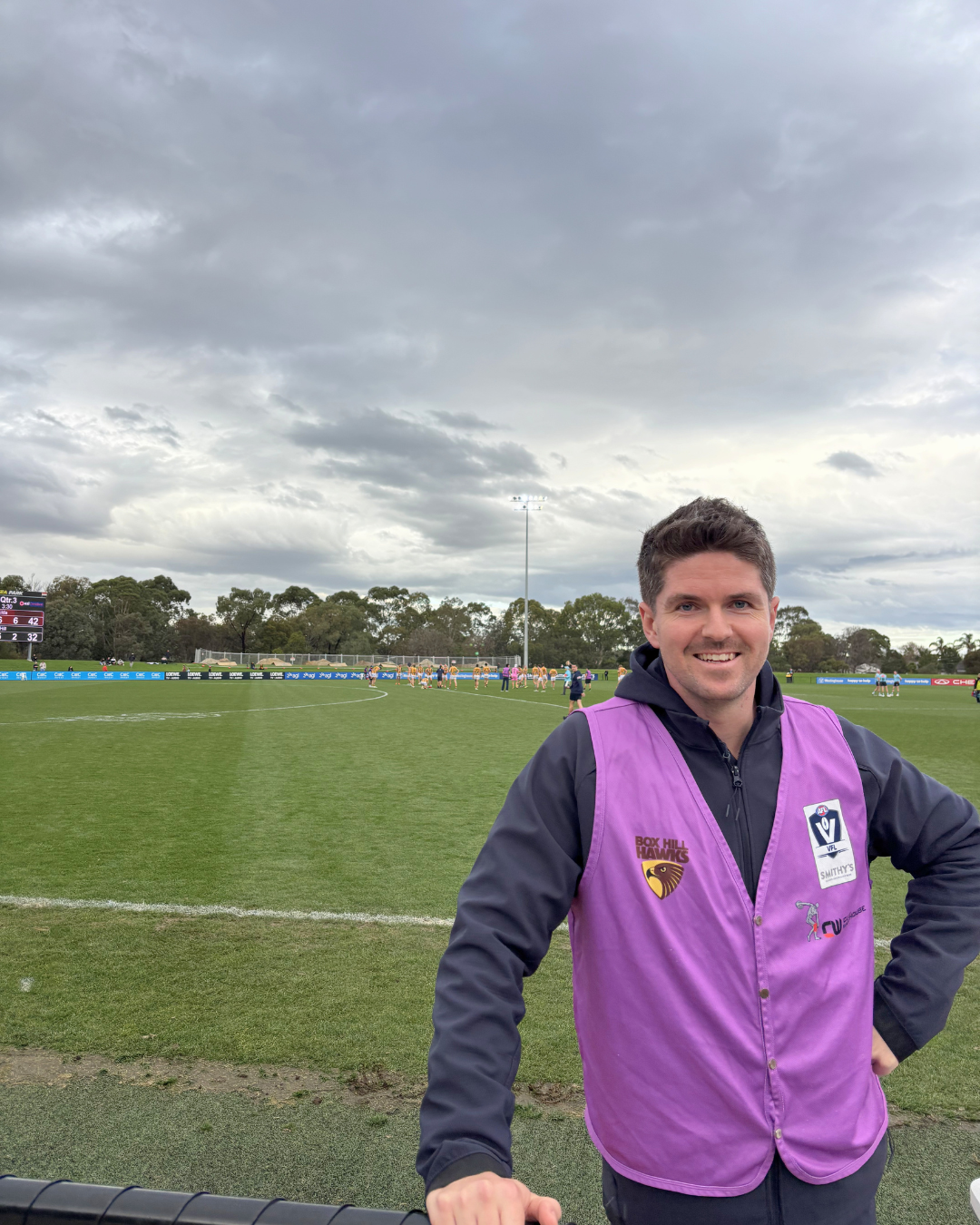 A man wearing a purple vest standing on a sports field during an Australian rules football game, with players and spectators in the background under a cloudy sky.