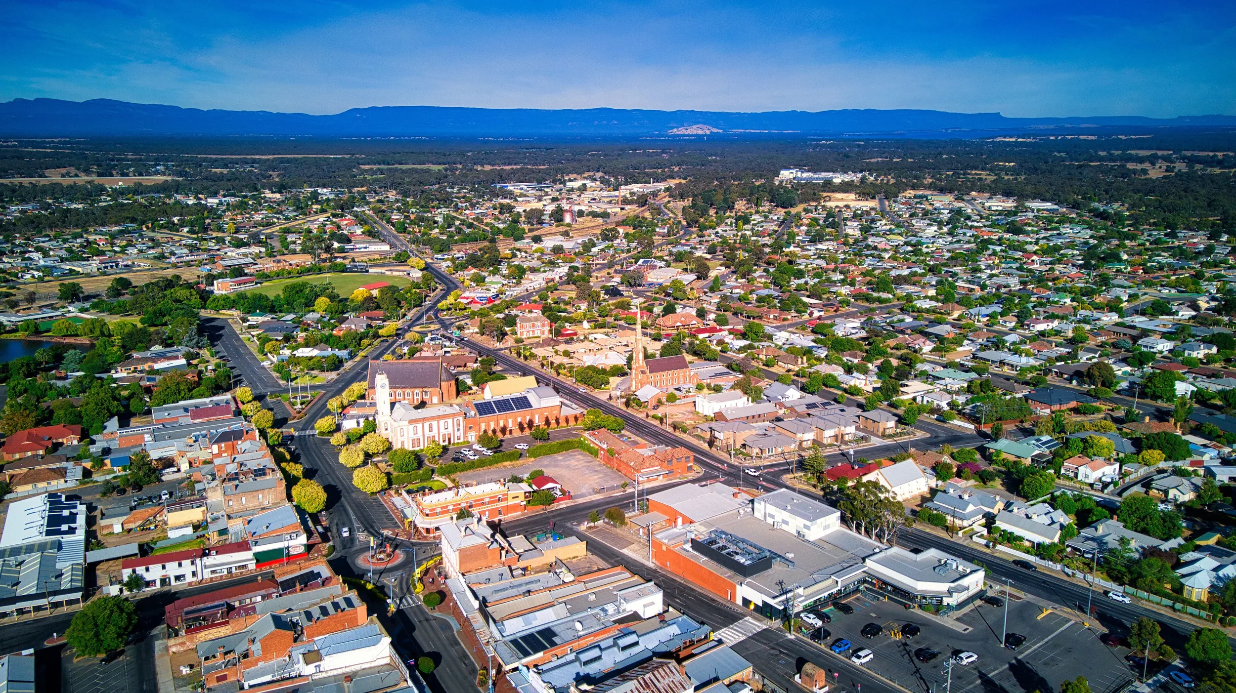 An aerial view of a small town with various buildings, trees, roads, and a church, with mountains in the background under a partly cloudy sky.