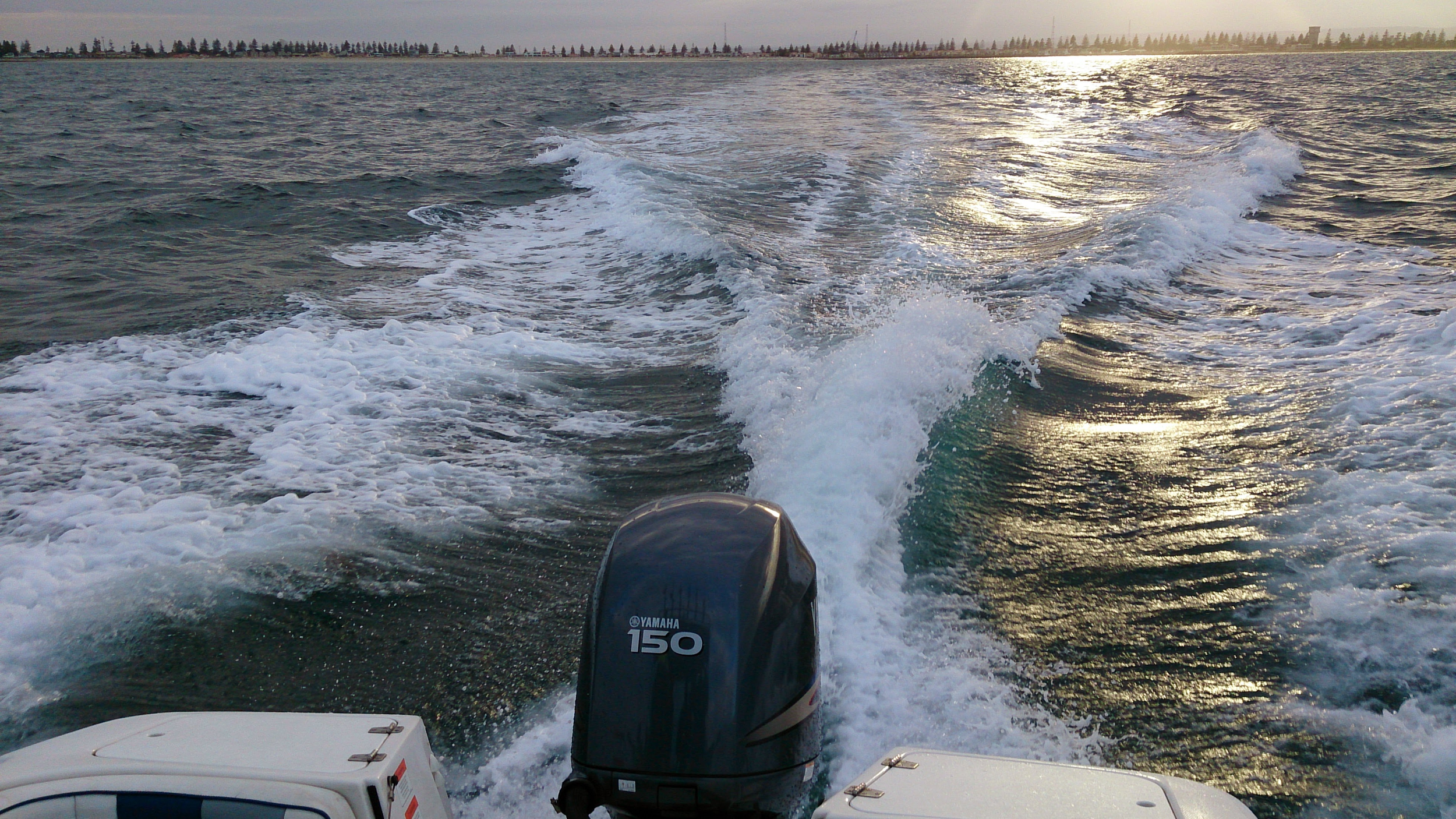View from a boat showing a Yamaha 150 outboard motor, churning water behind the boat, with a distant shoreline lined with trees and some structures, and the sun reflecting on the water's surface.