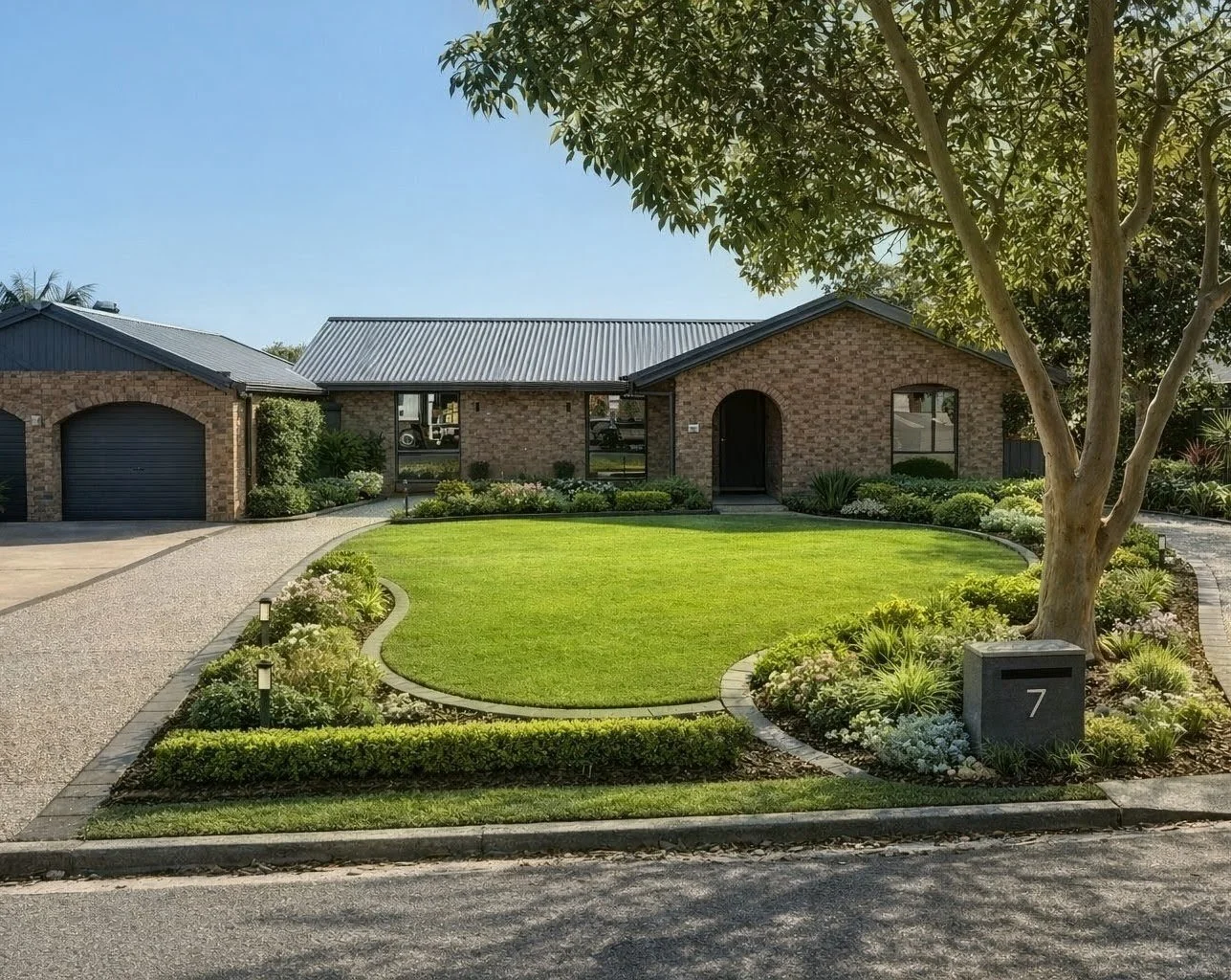Front view of a brick house with a well-maintained lawn, garden beds, a large tree, and a driveway.