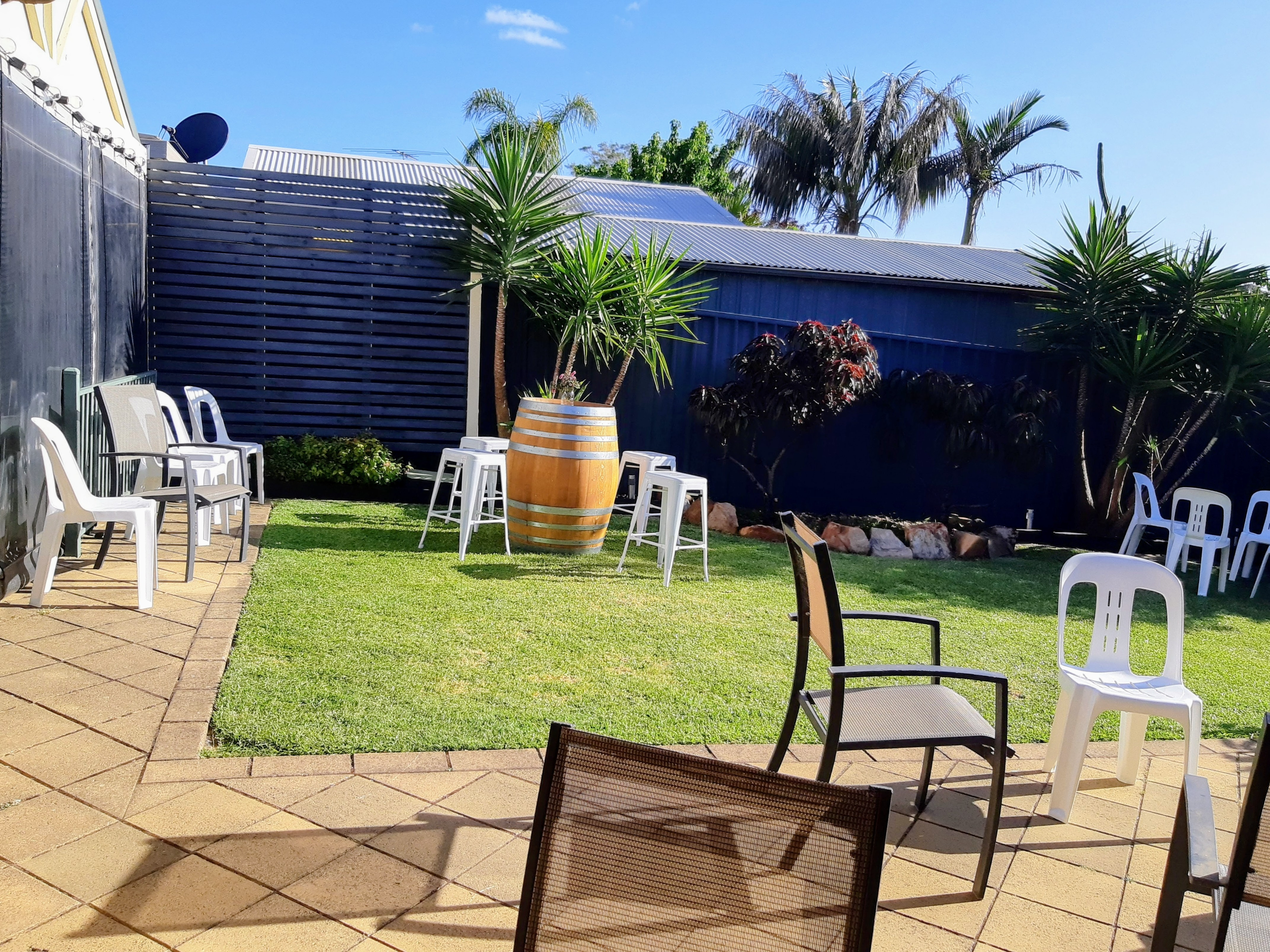 Outdoor patio area with chairs, tables, a barrel, a grassy patch, and several tropical plants and trees under a clear blue sky.