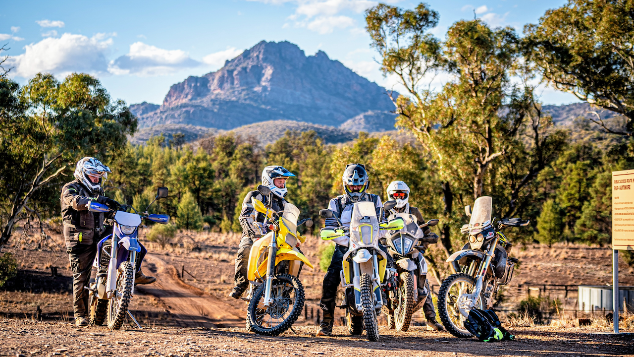 Five motorcyclists in riding gear and helmets with off-road motorcycles, standing on a dirt trail with trees and mountains in the background during daytime.