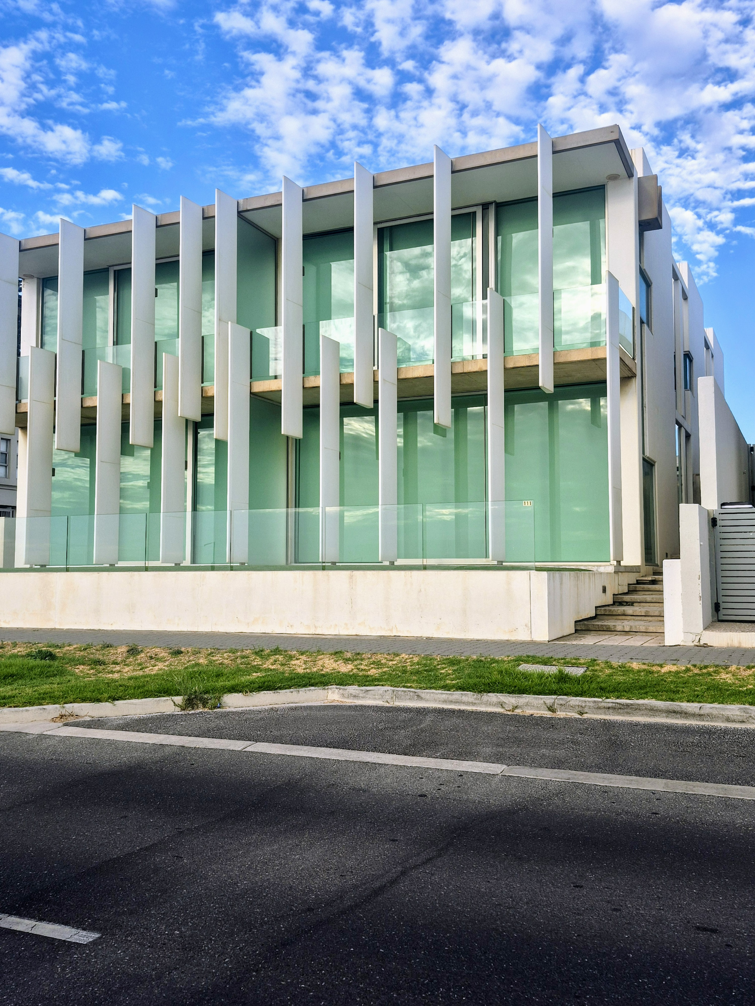 Modern multi-story building with large glass windows and white architectural accents, situated next to a parking lot with a grassy area in the foreground, under a blue sky with scattered clouds.