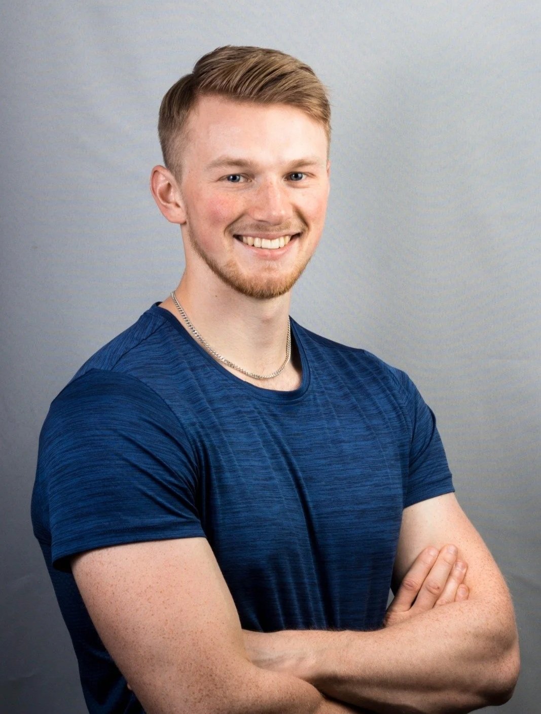 A young man with short blond hair, a goatee, smiling with arms crossed, wearing a blue athletic shirt and a silver chain necklace, standing against a plain gray background.