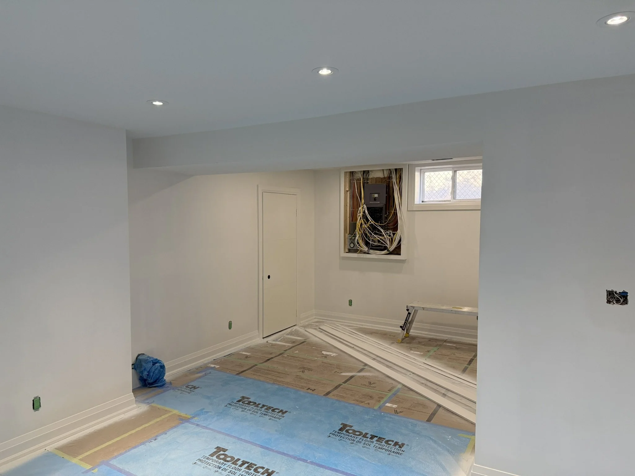 Interior room in a Mississauga basement under construction with white painted walls and ceiling, exposed electrical wiring, window, and protected flooring.