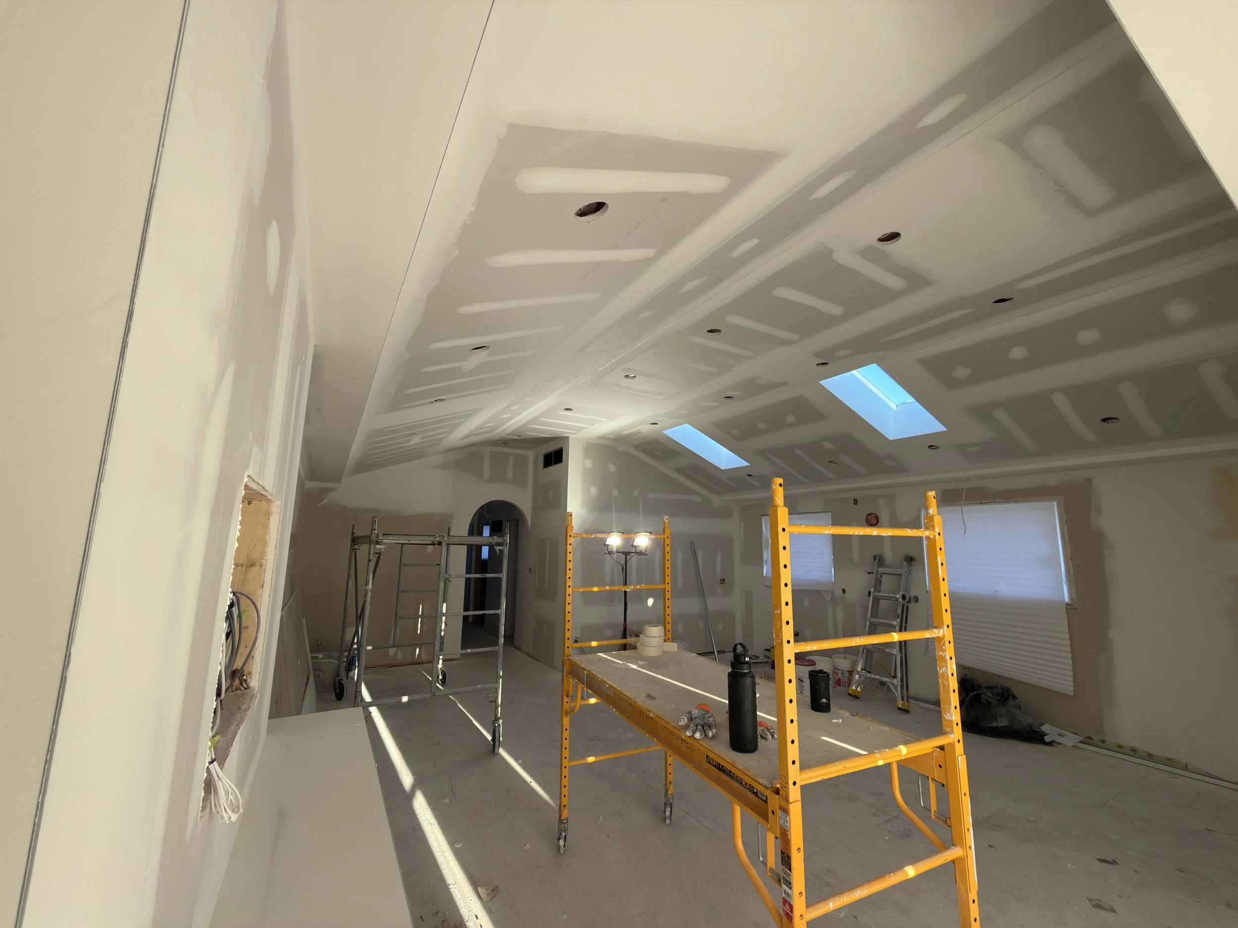 Interior of a room under construction with drywall ceiling and walls, scaffolding, and construction tools and materials.