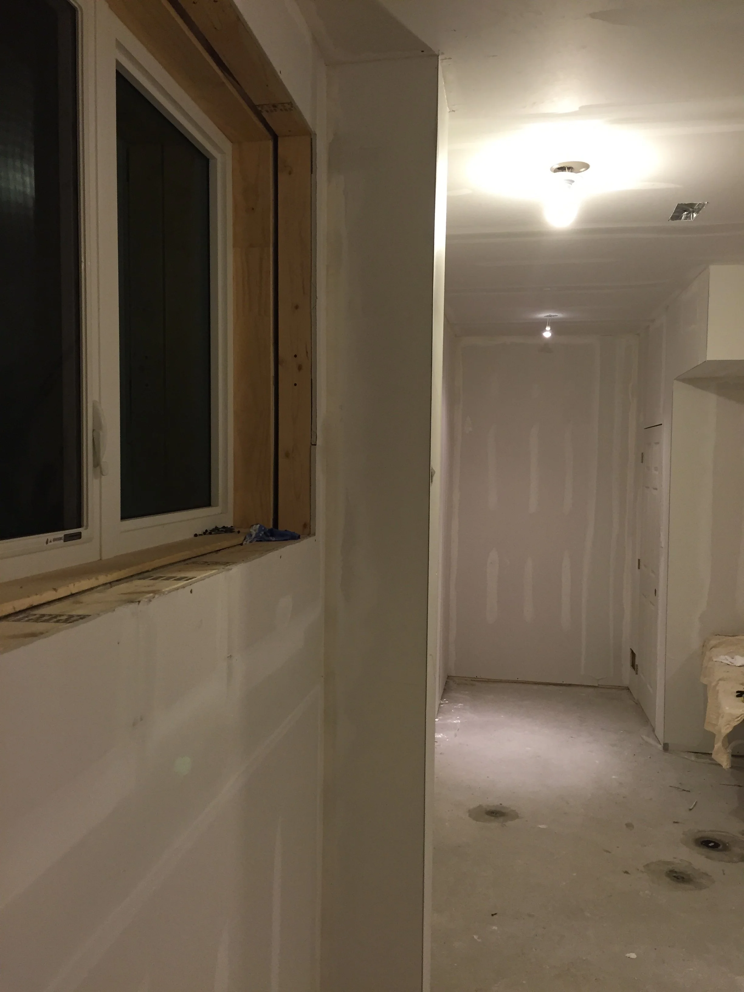 A hallway or corridor under construction, with unfinished drywall walls and ceiling, electrical outlets and recessed lighting, and some construction materials on the floor.