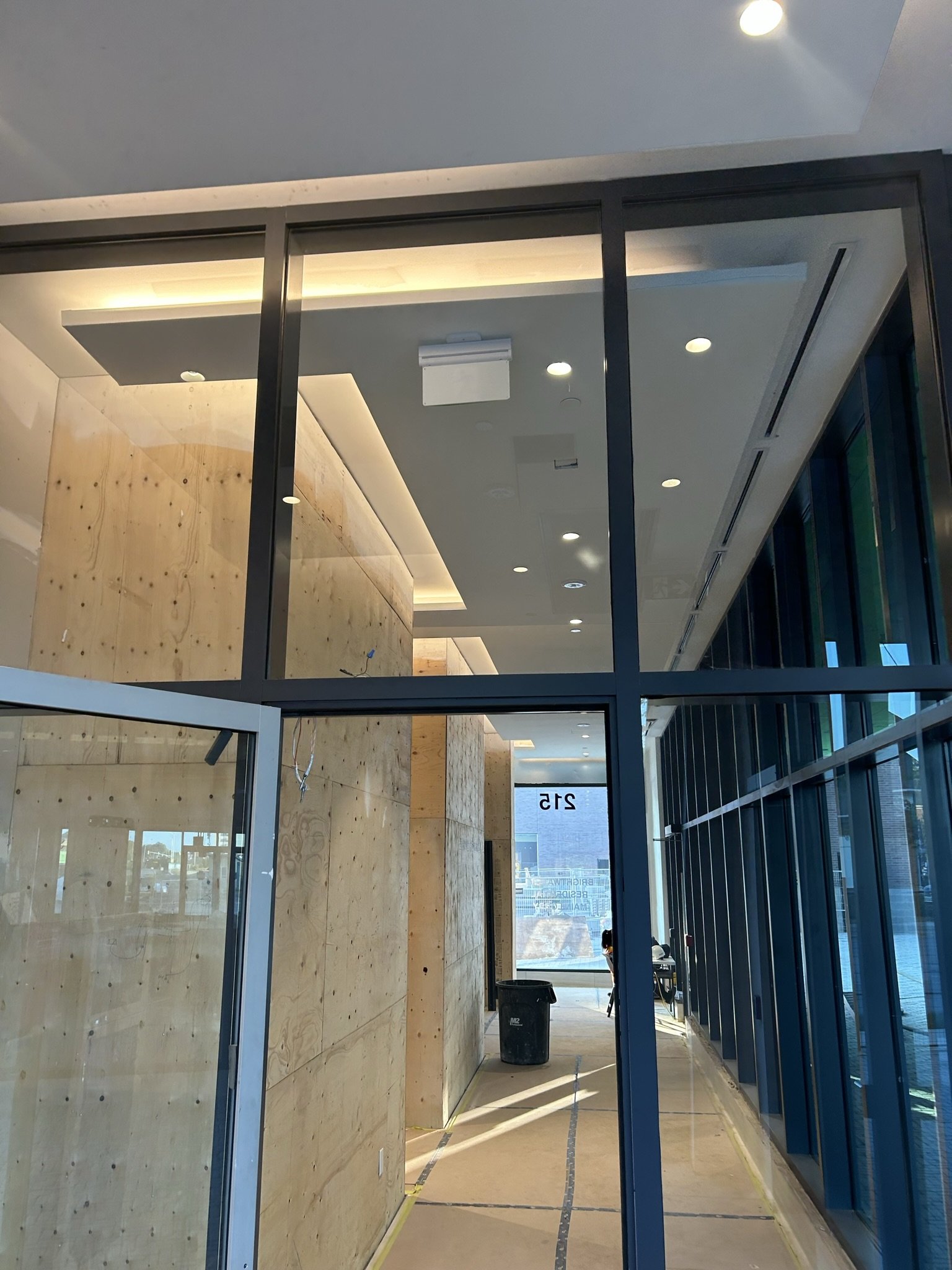 Interior of a building under construction with glass walls, wooden panels, and ceiling lights, showing a hallway leading outside.