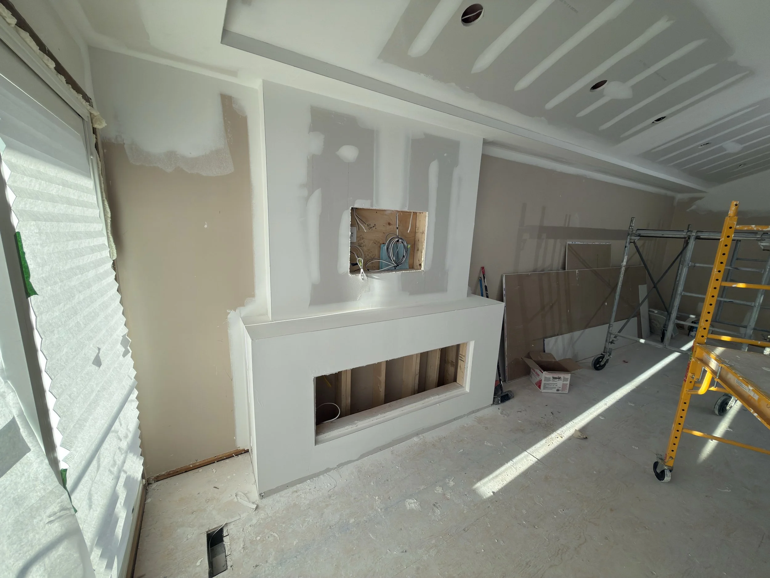 An unfinished room with drywall installed, some patches painted white, exposed electrical wiring in a cutout in the wall, a window with partially covered blinds, a yellow stepladder, and protective covering on the windows.