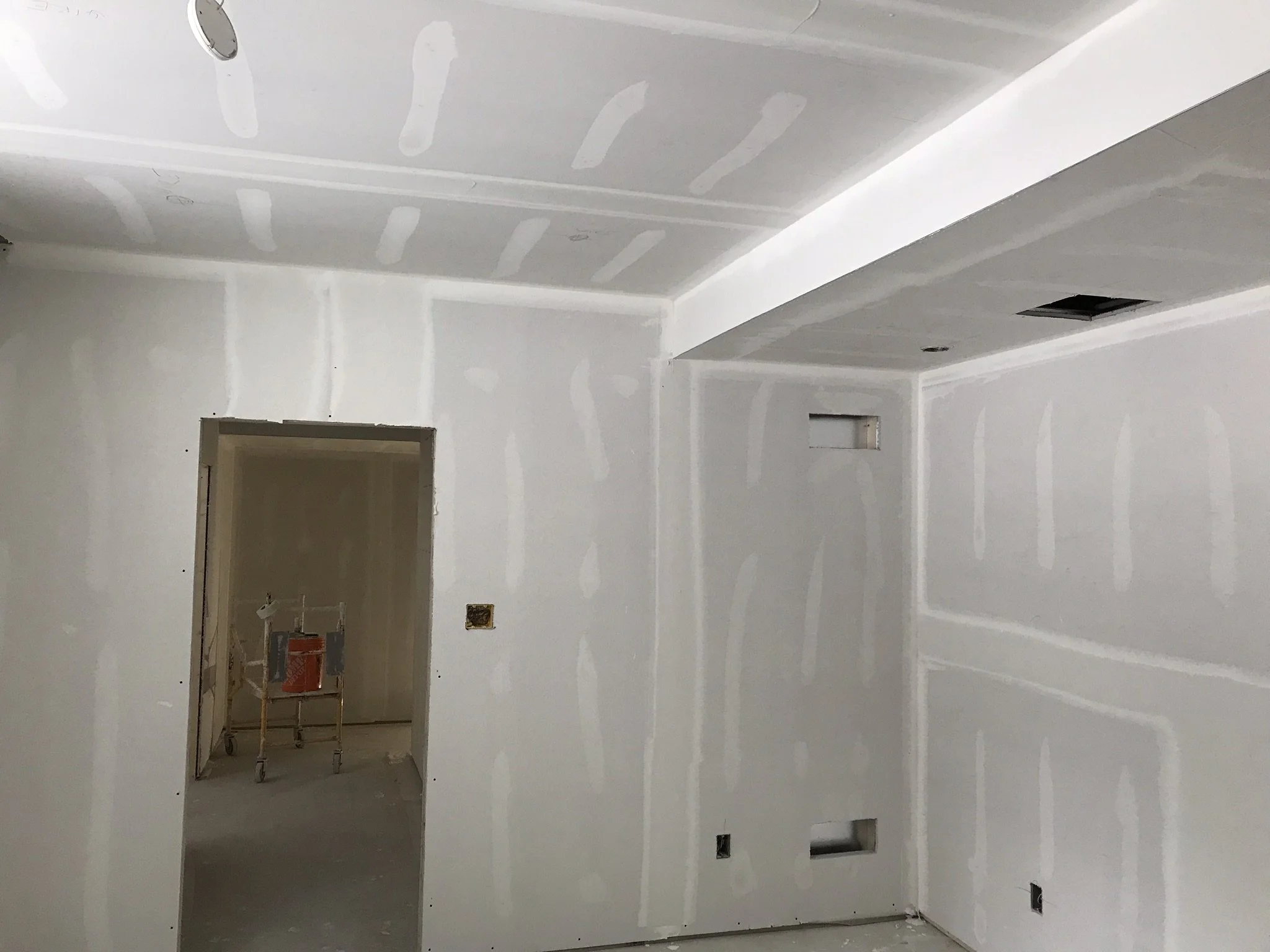 Interior room under construction with drywall installed on walls and ceiling, electrical outlets and wiring boxes visible, and some construction tools and materials present.
