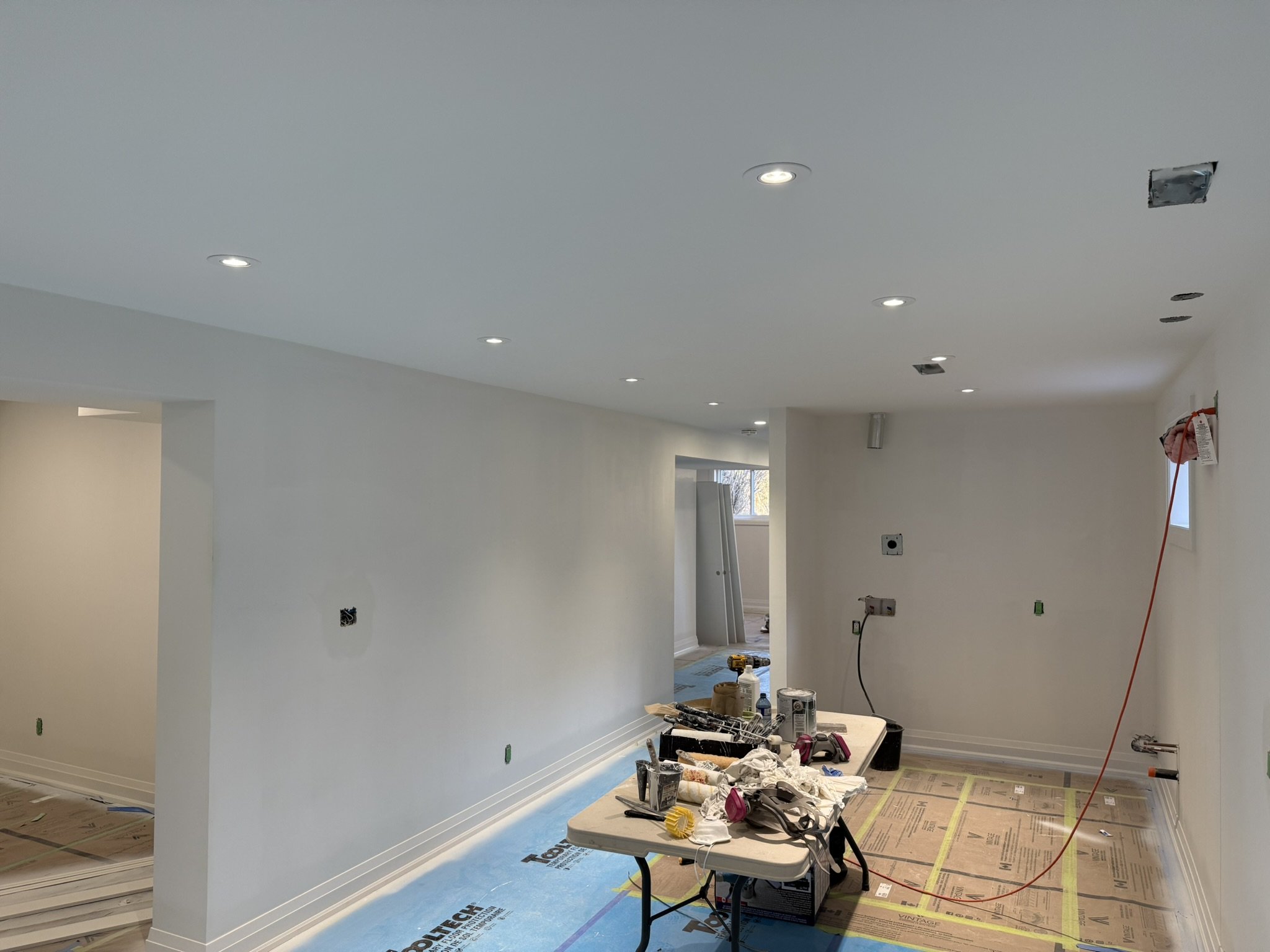 A basement room under renovation with a white ceiling fitted with multiple pot lights. The walls and ceiling are taped and painted white.