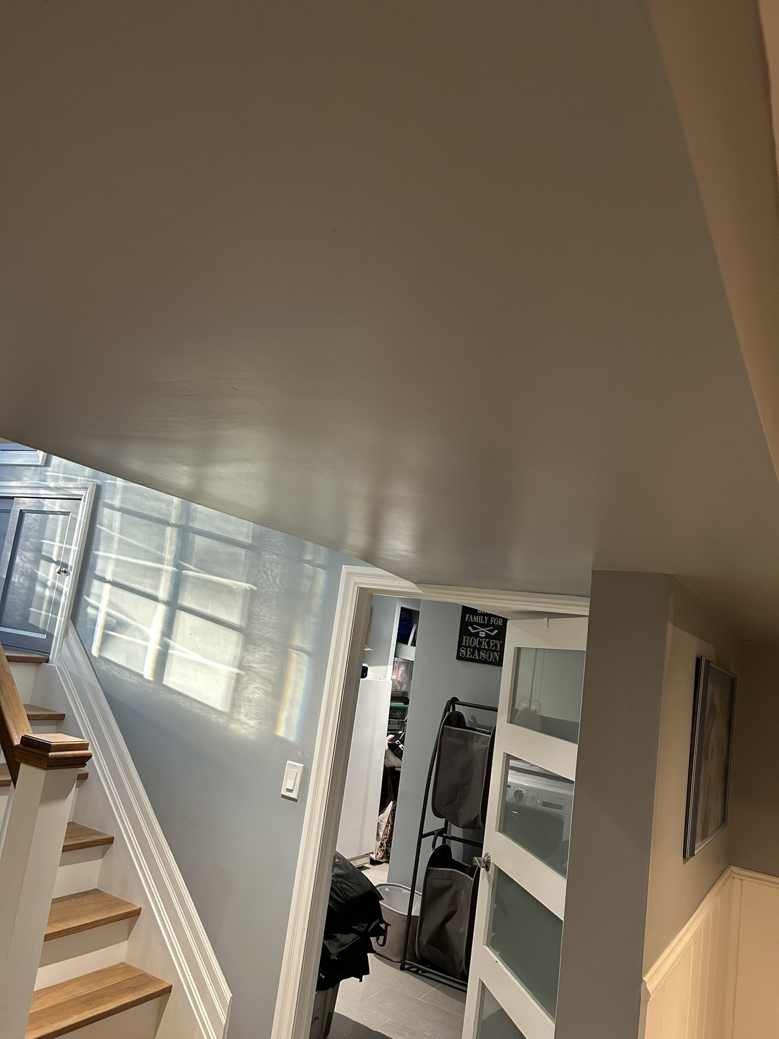 Interior view of a house with a staircase, a window casting sunlight inside, a white cabinet with glass doors, and various household items.