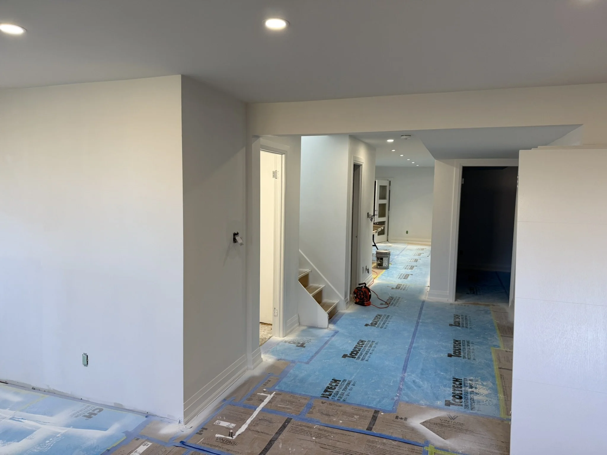 Interior of a house under construction with white walls, a blue tarp covering the floor, and recessed lighting on the ceiling.