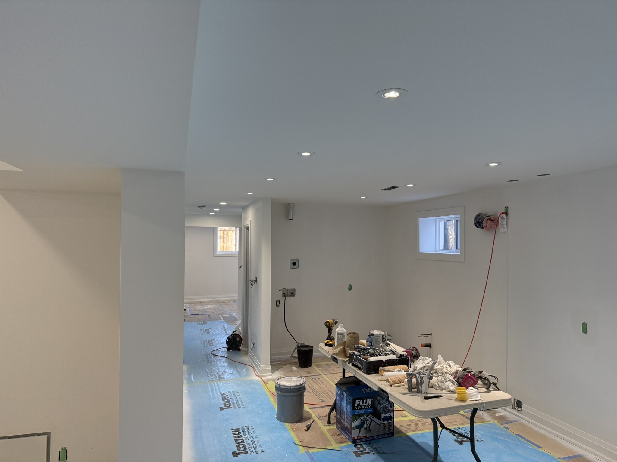 Interior of a room under renovation with ceiling lights installed, construction tools, and supplies on a table and floor, windows providing natural light, and walls painted white.