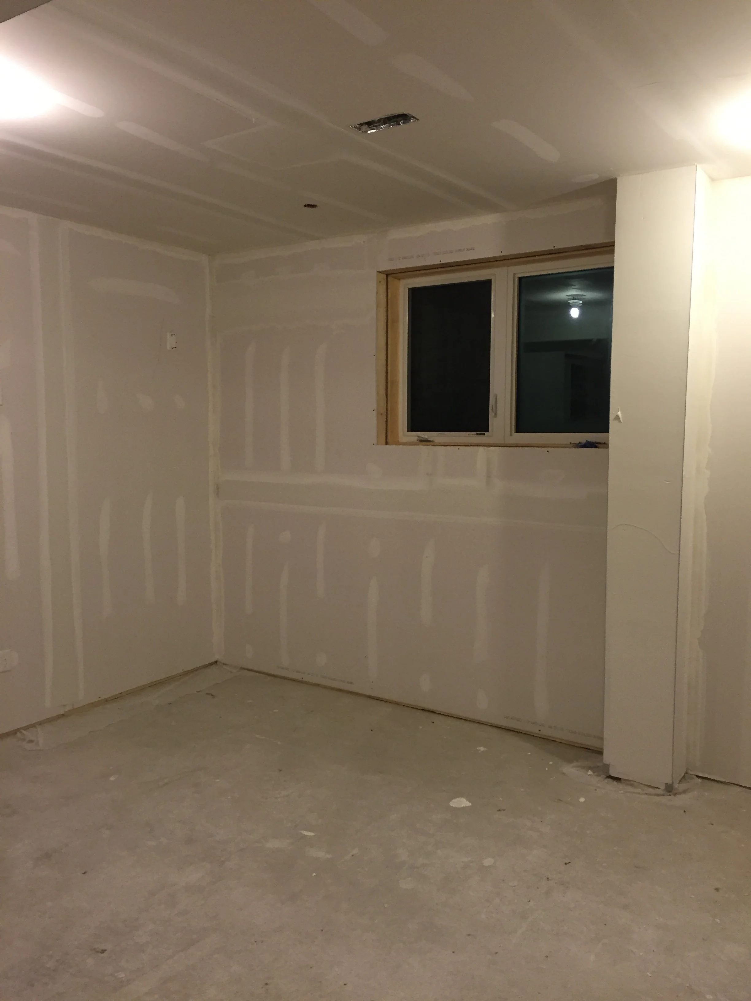 Unfinished room with drywall installed, visible seams, and no flooring. A window is on the wall, revealing a dark exterior view.