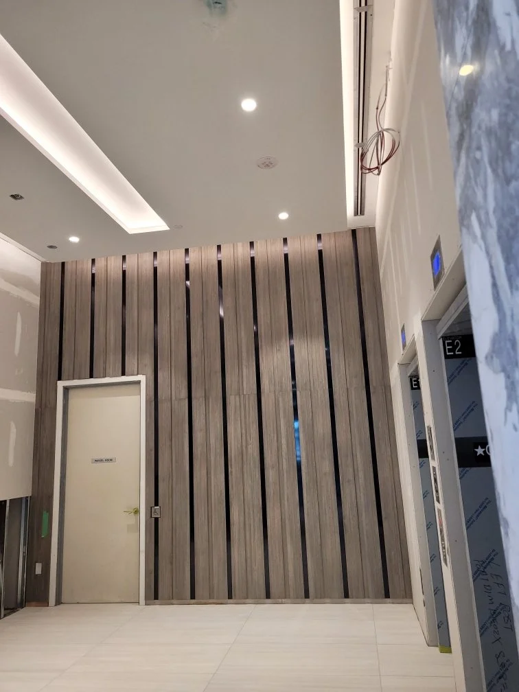 Interior view of a modern building lobby with elevator doors, a wood-paneled wall, a door labeled 'Meeting Room,' and a ceiling with recessed lighting.