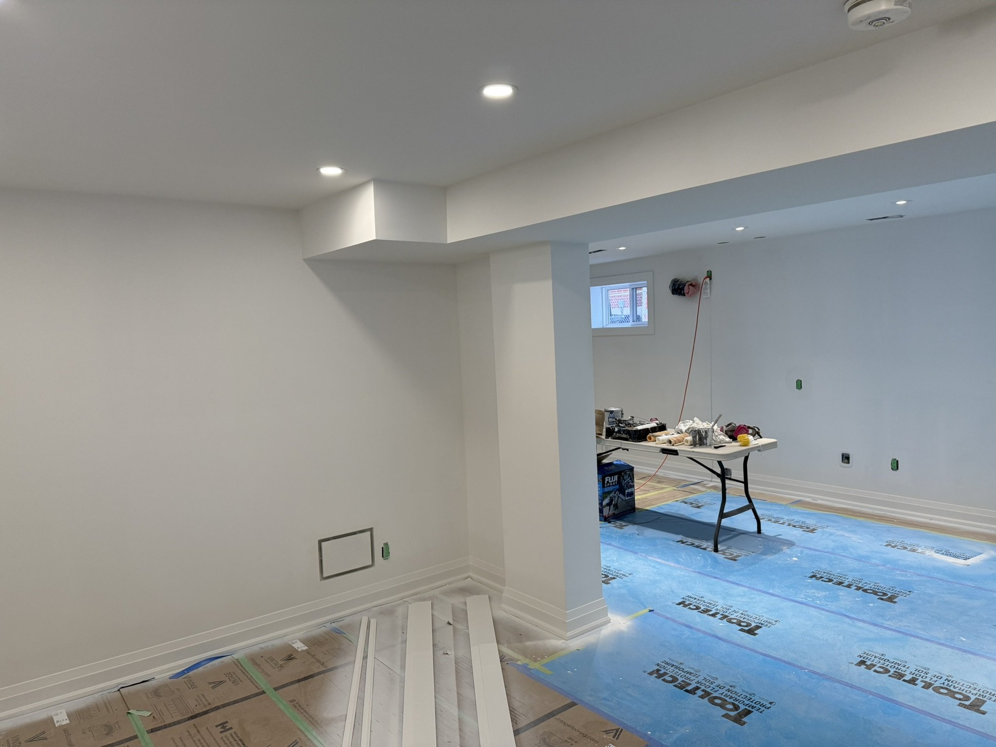 Interior of a room under construction or renovation, with white walls and a drop ceiling with recessed lighting. A small window is visible, and there is a table with tools and materials. Blue protective covering is on the floor, and sheets of paper o
