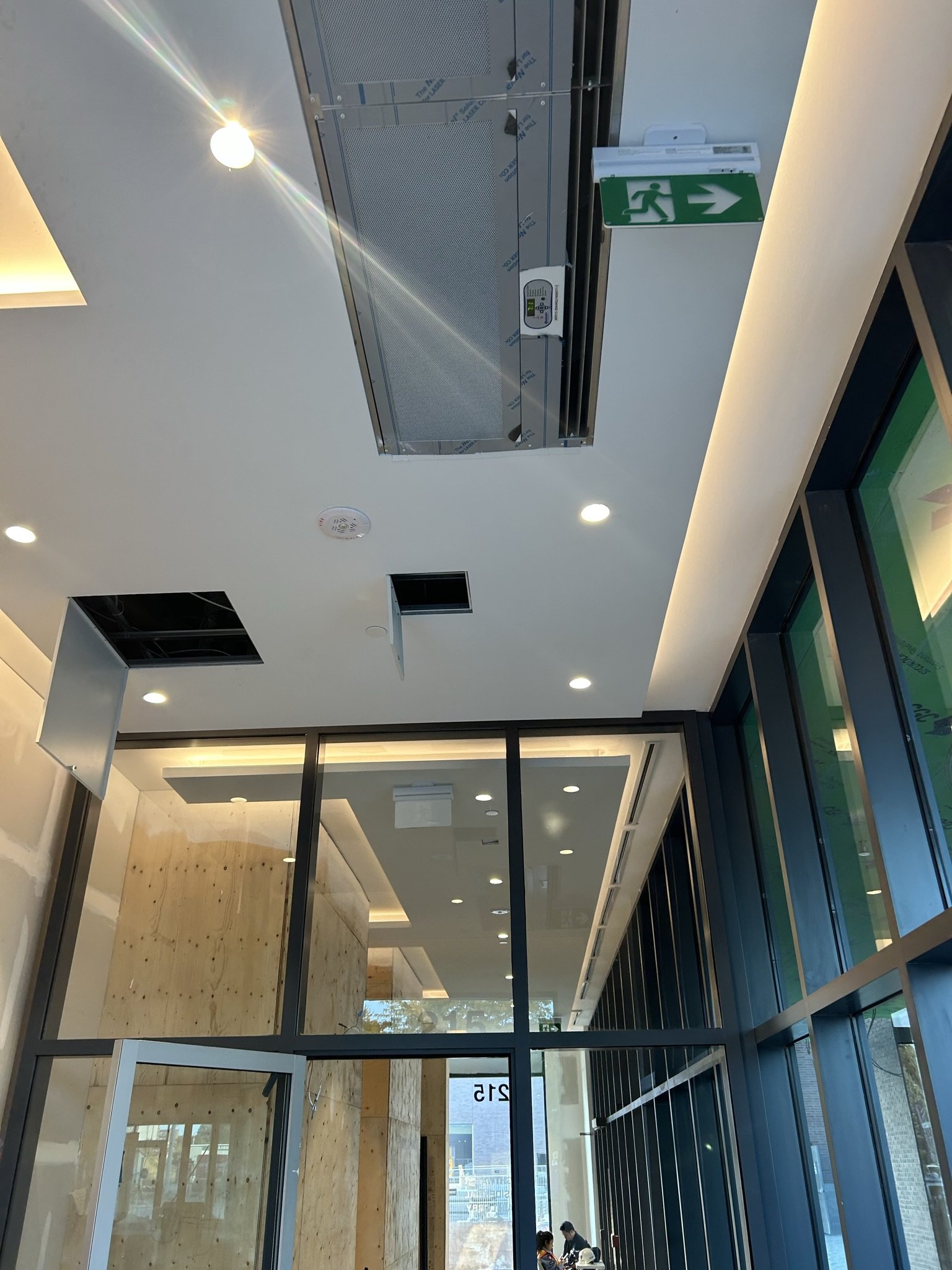 Interior view of a modern building with glass walls, ceiling lights, an industrial air conditioning unit, and a green exit sign with a running person and arrow.