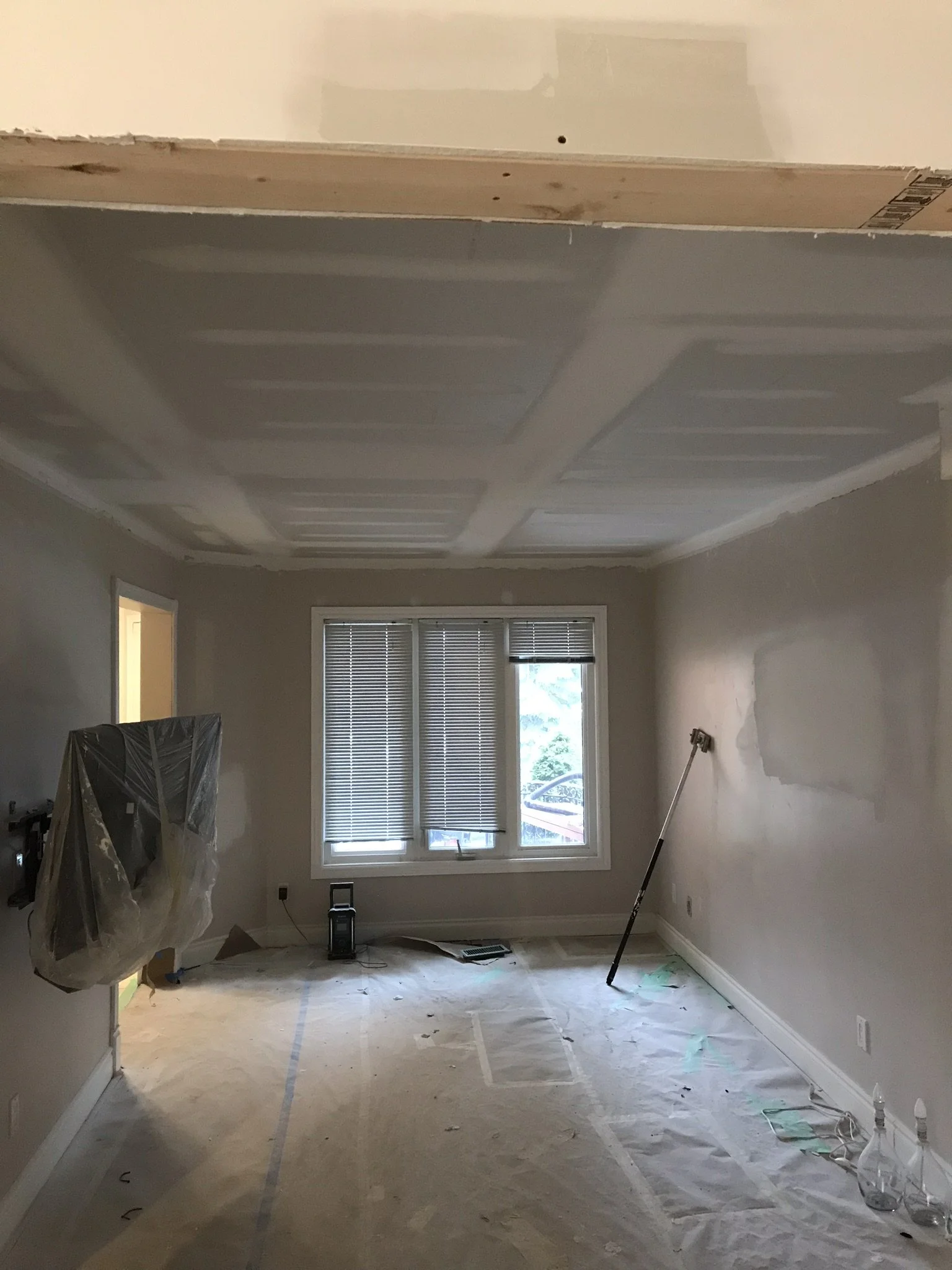 A room under renovation with an unfinished ceiling and walls. The floor is covered with protective paper, and there are painting supplies, tools, and equipment scattered around, indicating ongoing work.
