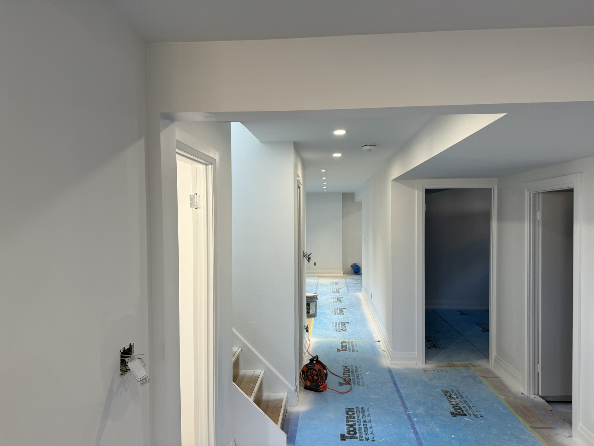 A hallway under construction with white walls, multiple ceiling recessed lights, and protective floor covering. The hallway leads to several doorways, some of which are open, showing adjoining rooms, with tools and construction materials visible.