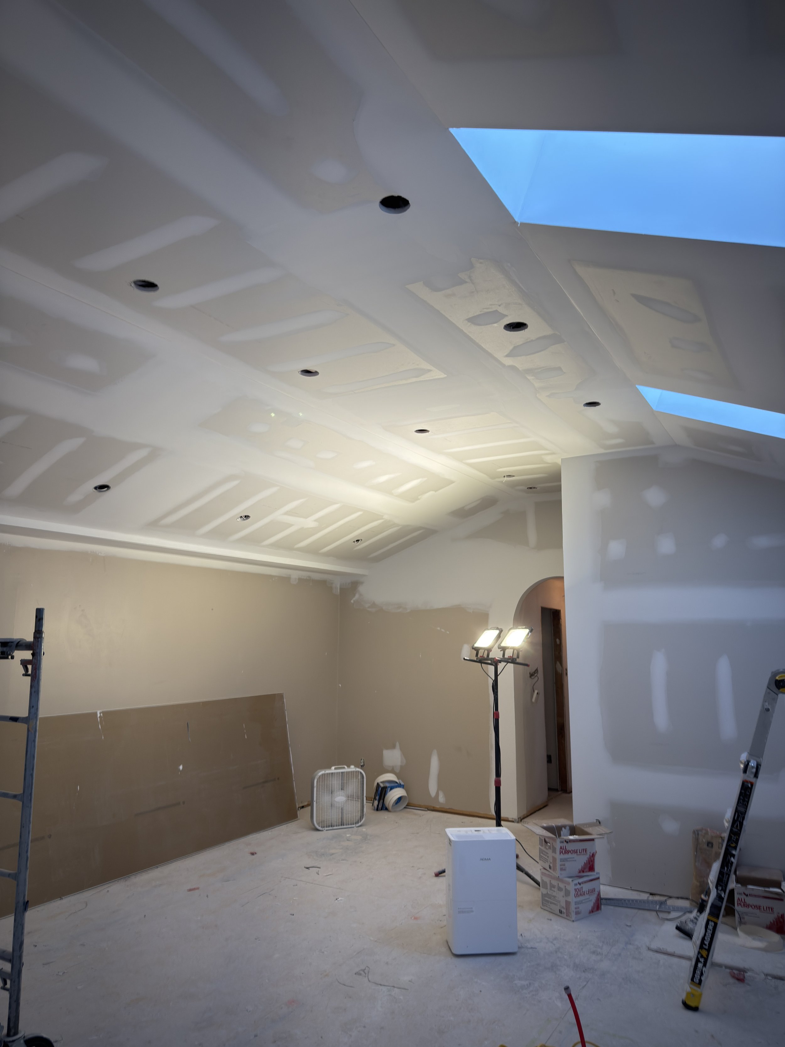 Ceiling and wall drywall installation in a room under construction, with construction tools, boxes, and lighting equipment present.