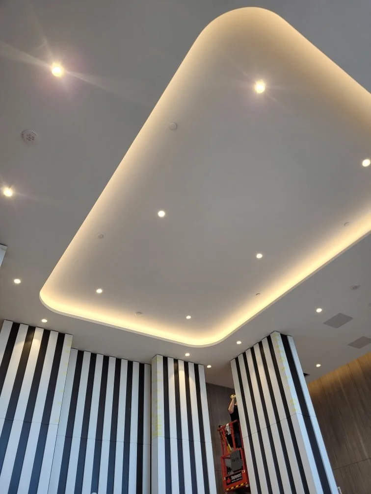 Interior ceiling with recessed lighting and a curved, illuminated soffit above striped black and white walls, with a worker on a lift.