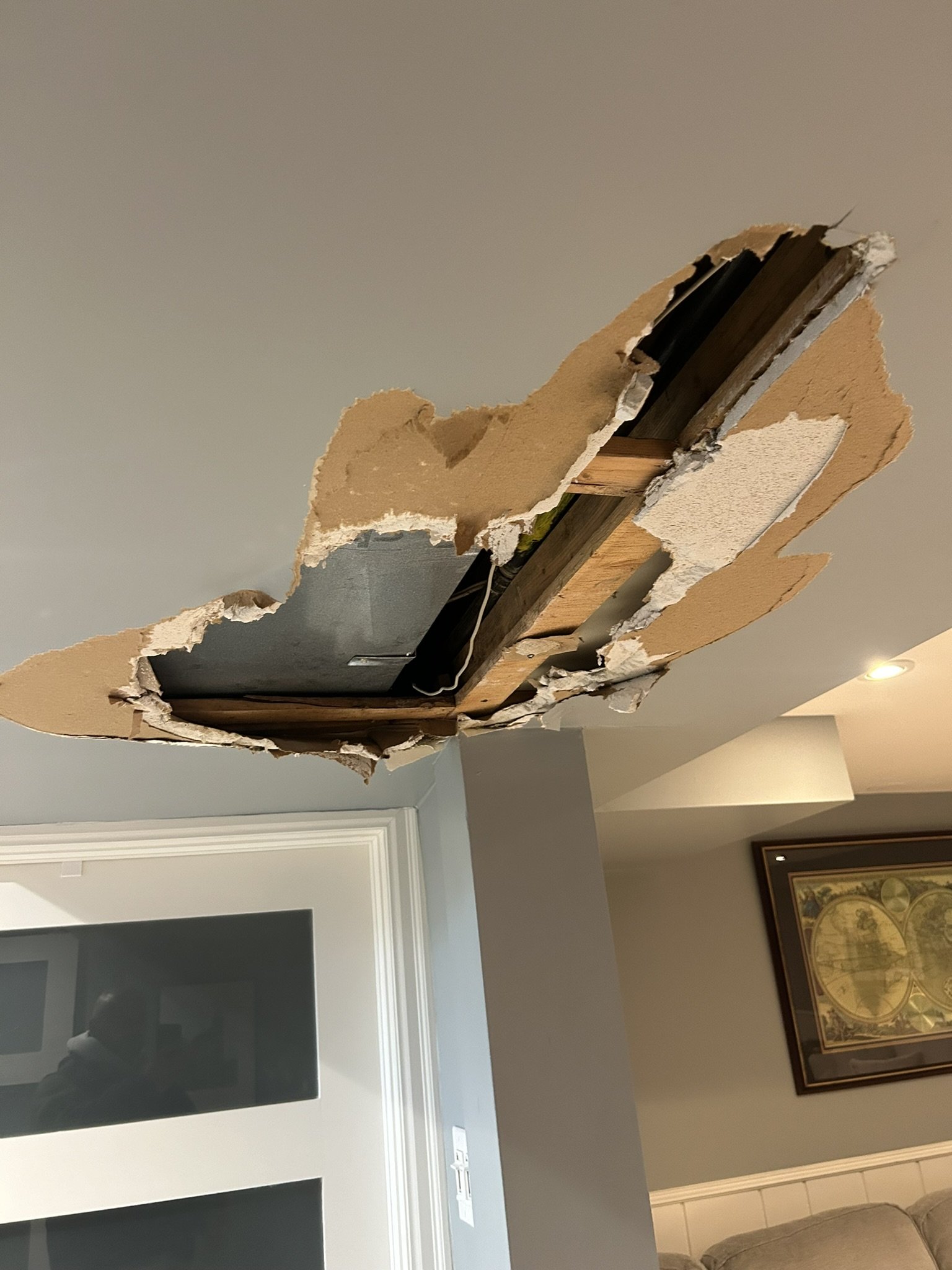 A ceiling with a large hole revealing wooden beams and black wires, with a framed map on the wall and a beige sofa in the room.