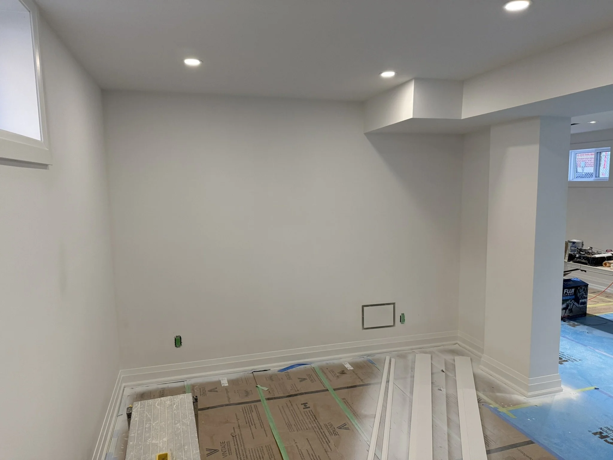A room under renovation with white walls, recessed lighting, a window, and construction materials on the floor and in the background.