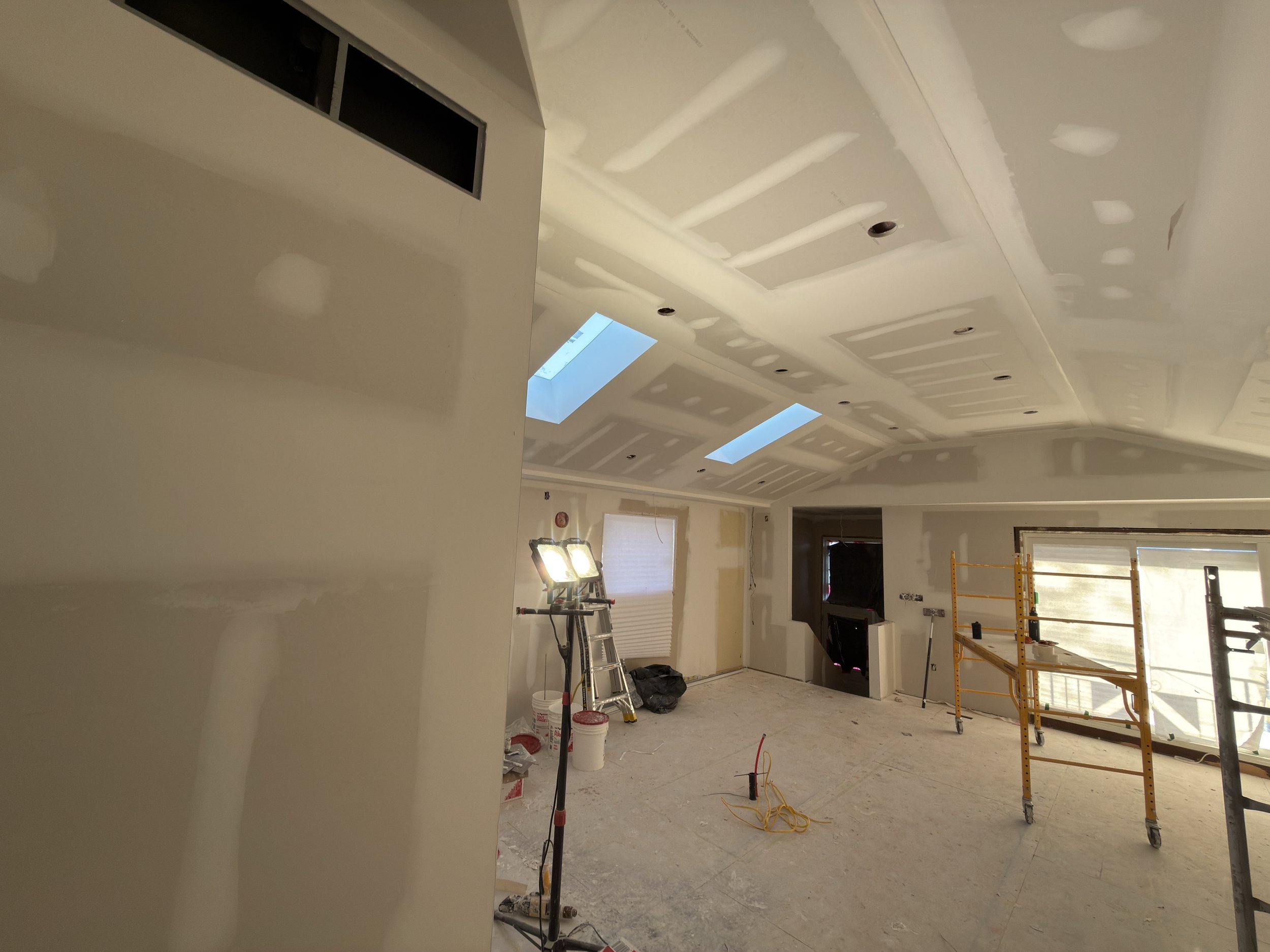 Interior room under renovation with unfinished drywall ceiling, construction tools, ladders, and scaffolding. Light is coming through skylights and windows.