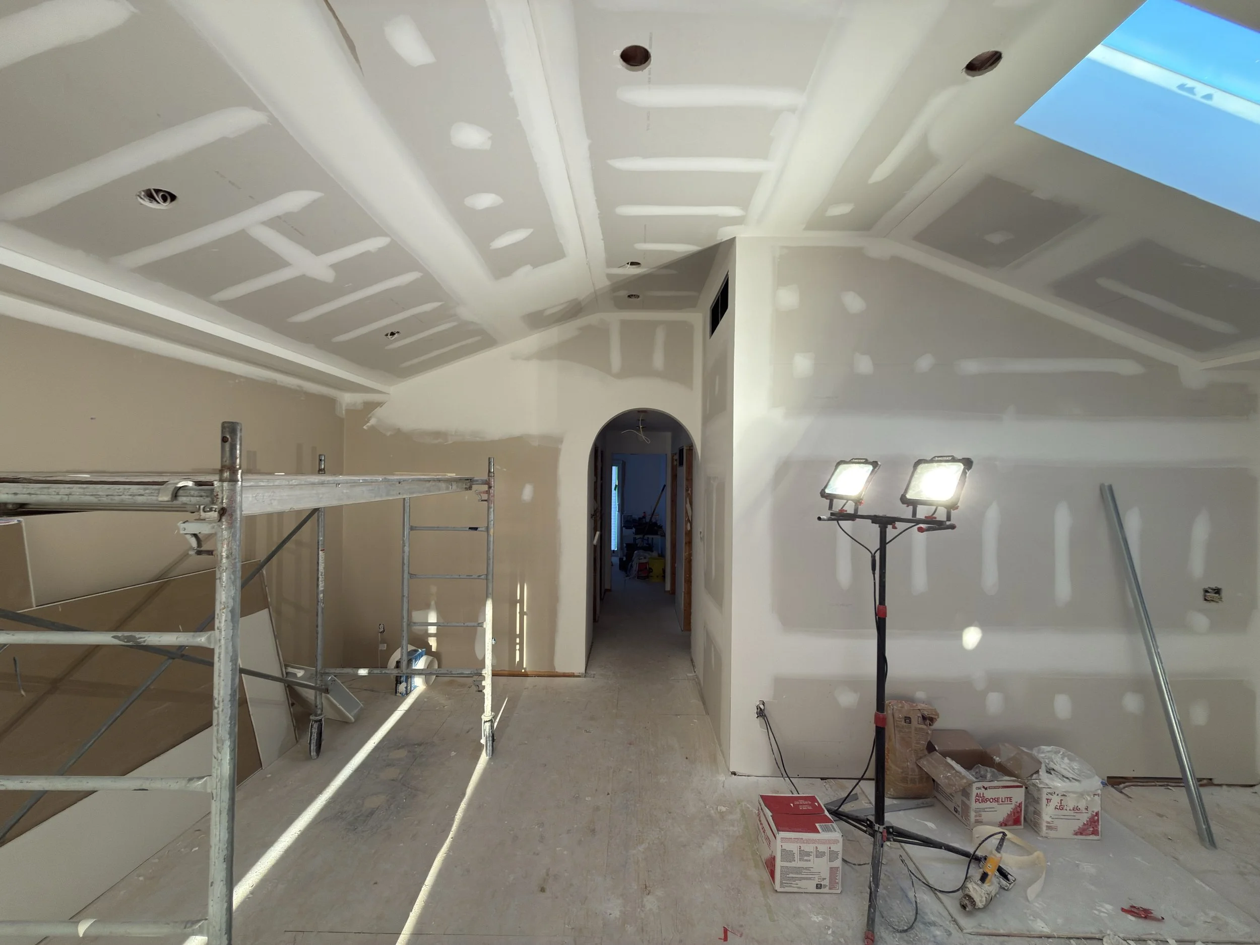 Interior of a house under construction with drywall ceiling and walls, scaffolding, construction lights, and tools and materials on the floor.