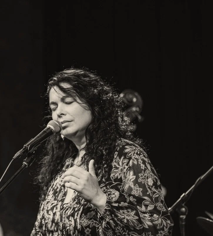 A woman with curly hair singing into a microphone on stage, with closed eyes and a hand placed on her chest.