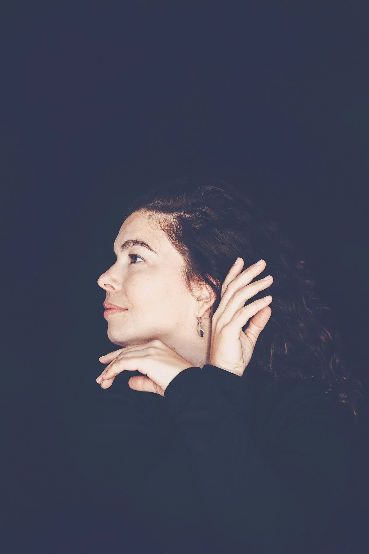 A woman with curly dark hair poses against a dark background, resting her chin on her hand and holding her ear with her other hand, looking to the side with a slight smile.