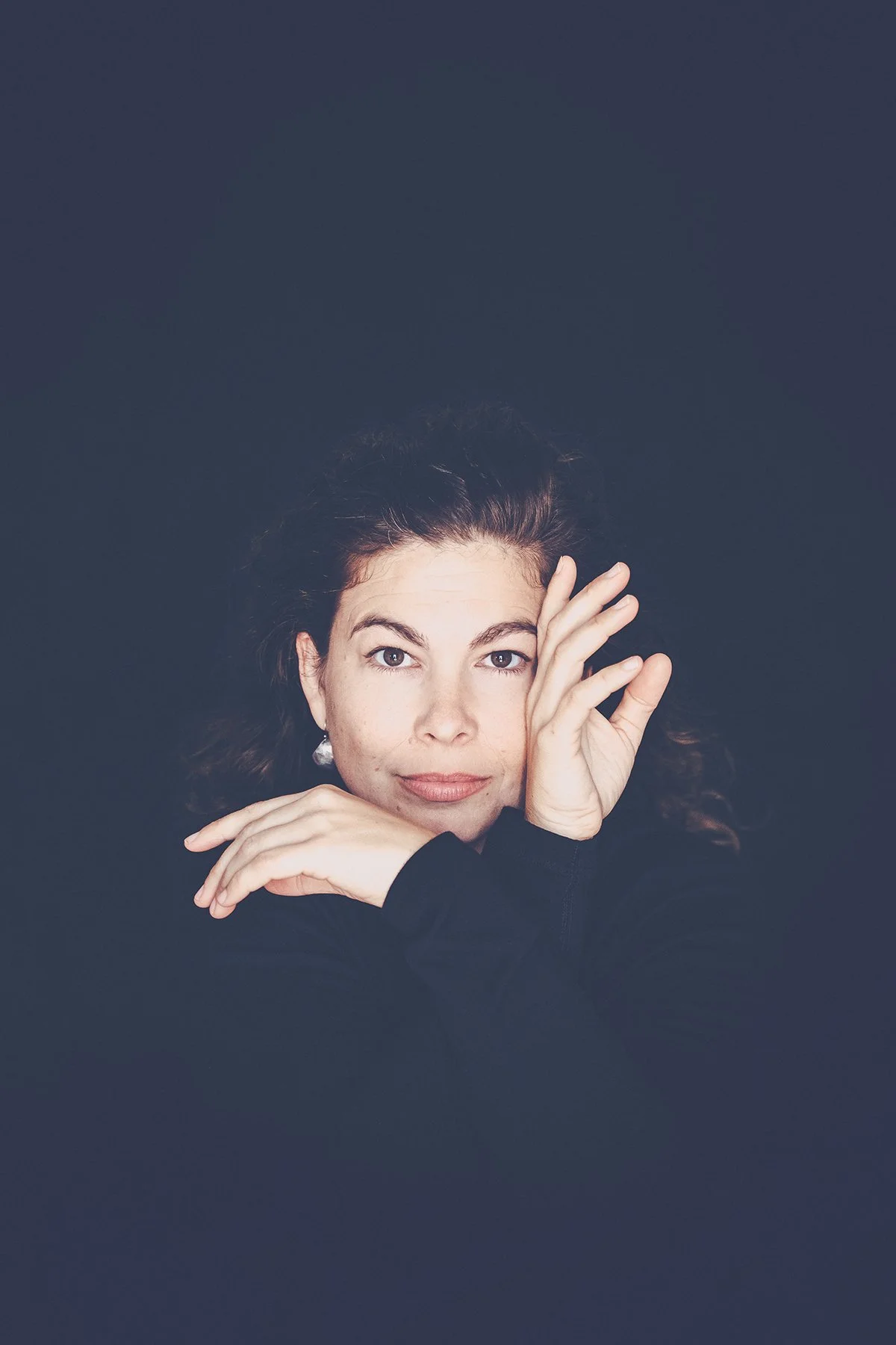 A woman with light skin and dark, curly hair posing with her right hand touching her face and her left arm bent, resting her chin on her hand, against a dark background.