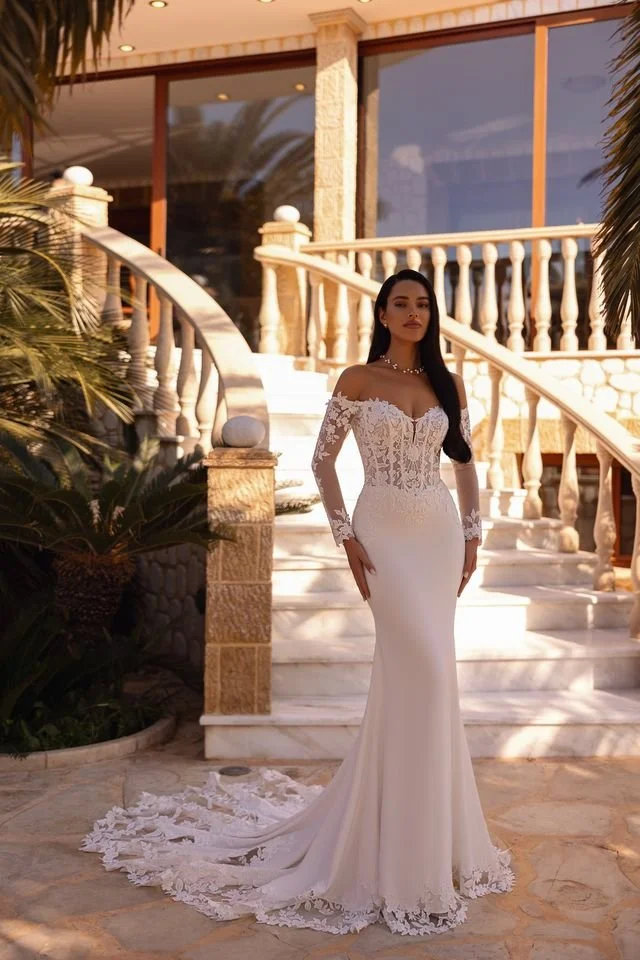 Brunette woman in white wedding dress with lace details standing outside on marble stairs in front of a villa with glass windows, surrounded by palm trees.