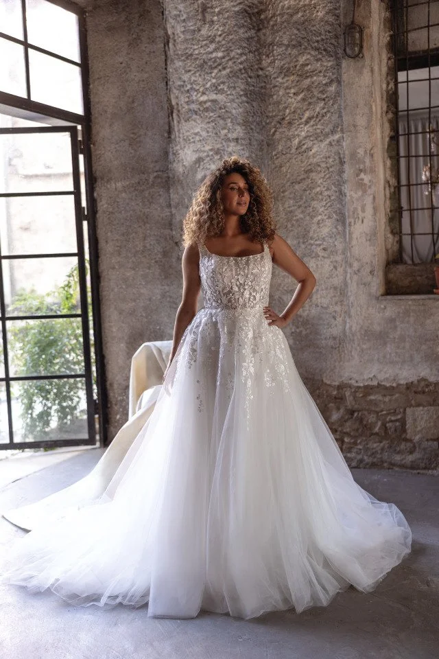 Woman wearing a white wedding dress with lace bodice and tulle skirt standing in an industrial-style room with exposed brick walls and large windows.
