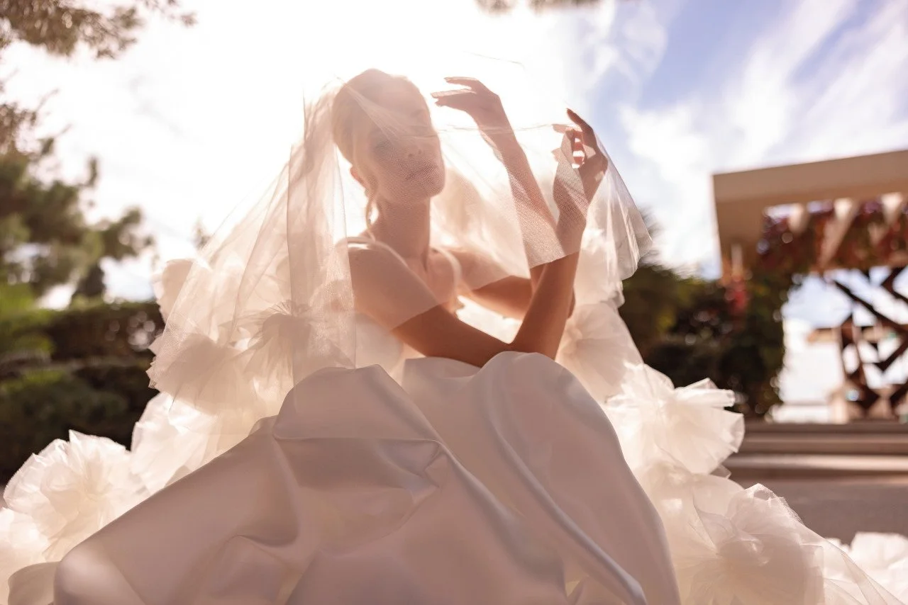A woman in a wedding dress and veil sits outdoors, backlit by the sun, with trees and a wooden structure in the background.