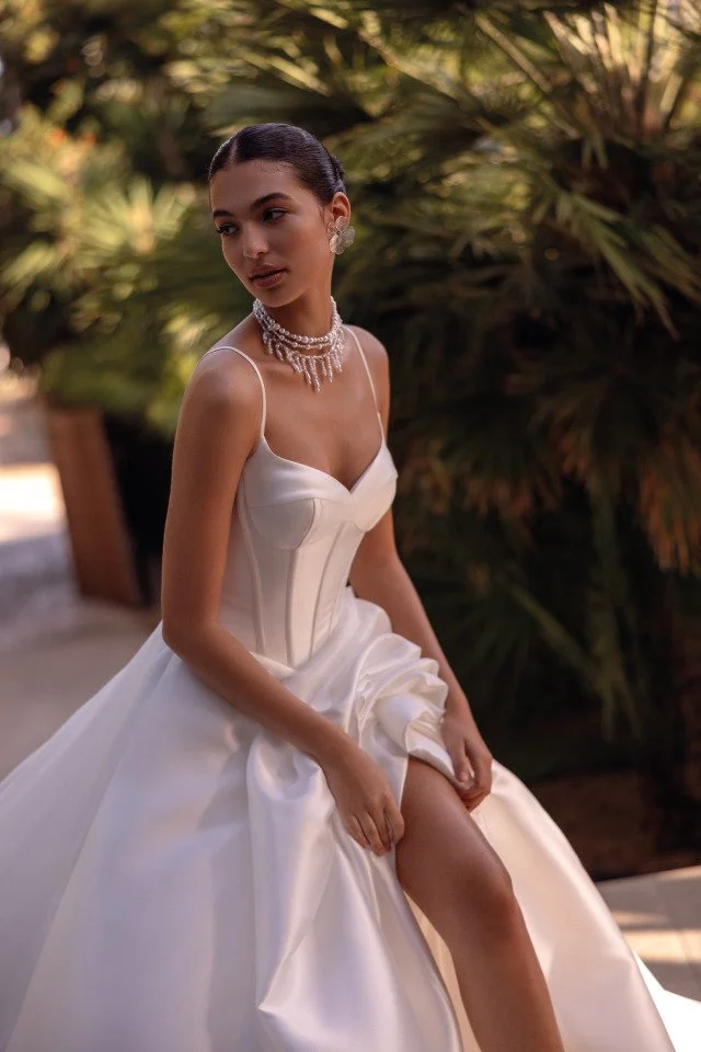 A woman in a white wedding gown with a fitted bodice and full skirt, sitting outdoors among green foliage, adjusting her gown's slit.