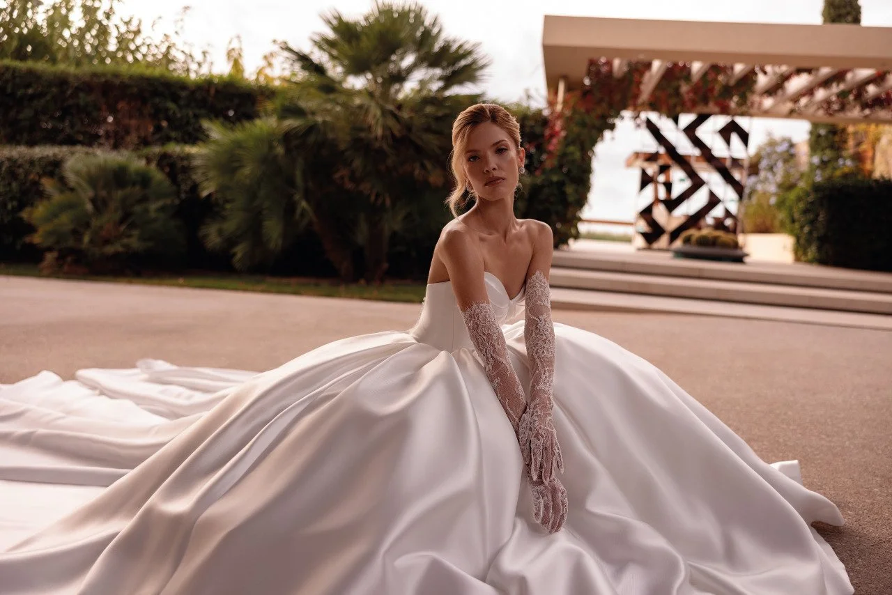 A woman in a white wedding dress sitting on the ground outdoors with green foliage and modern architectural elements in the background.