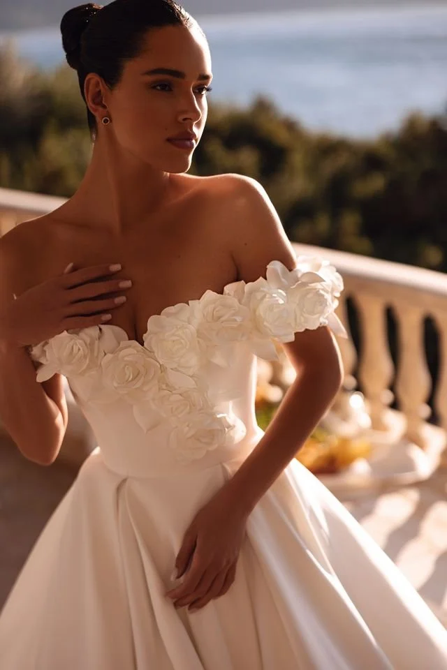 Woman in a white wedding dress with floral embellishments, standing outdoors on a balcony, with a blurred scenic background.