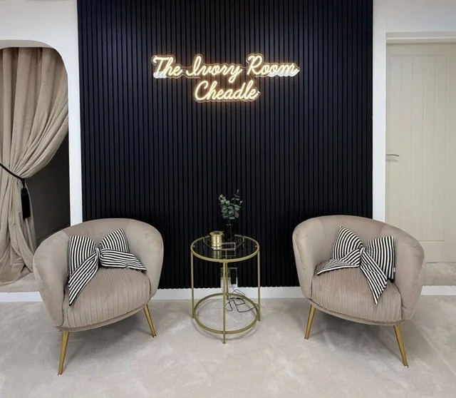 Two beige armchairs with striped pillows, a small round gold side table with a plant, against a dark panel wall with a neon sign reading "The Ivory Room Cheadle."