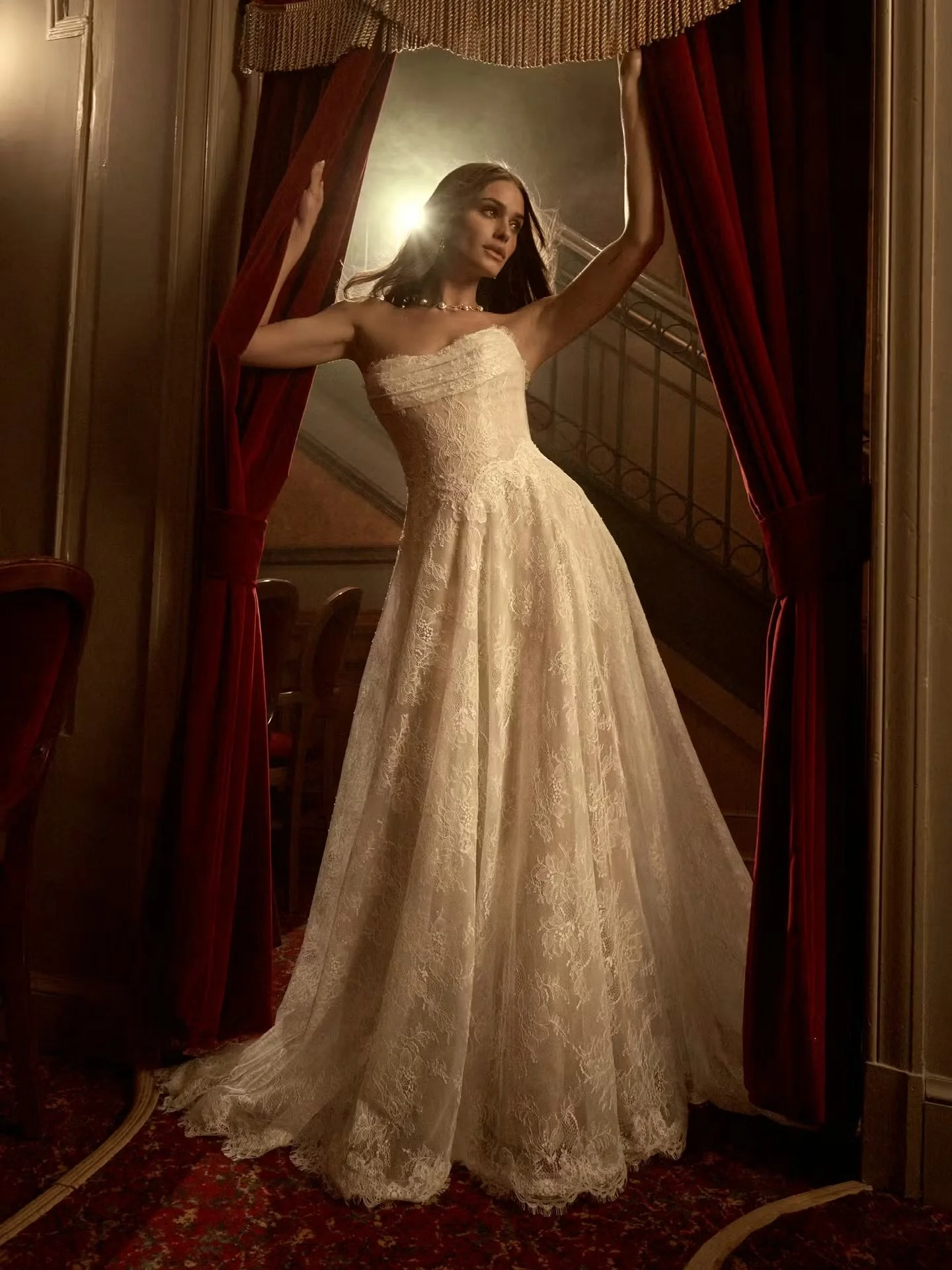 A woman in a wedding dress standing behind red velvet curtains, holding them open with both hands, in a dimly lit room with wood-paneled walls and a staircase in the background.