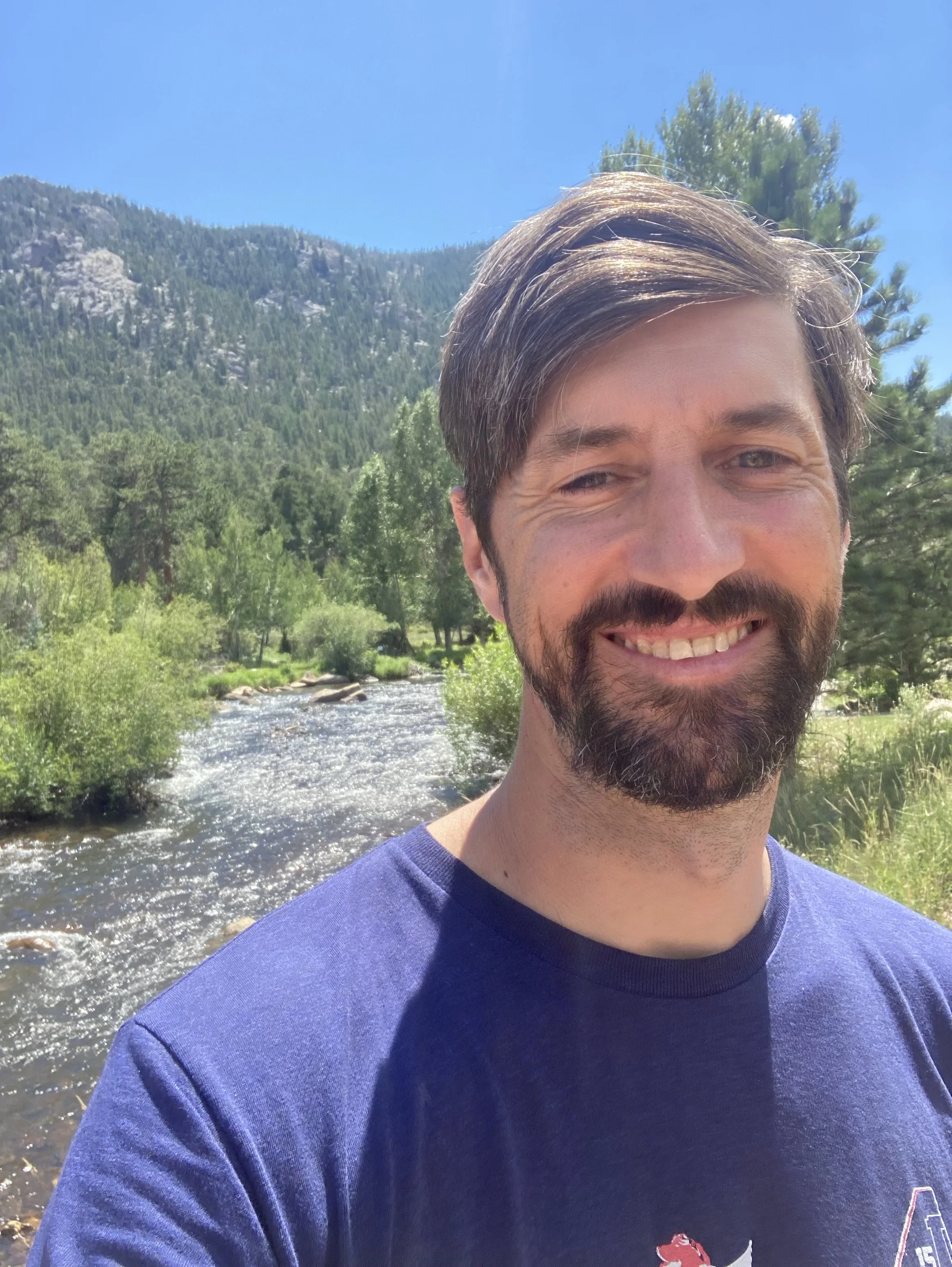 Close-up of a smiling man with brown hair and a beard, wearing a blue T-shirt, outdoors near a river with trees, mountains, and a blue sky in the background.
