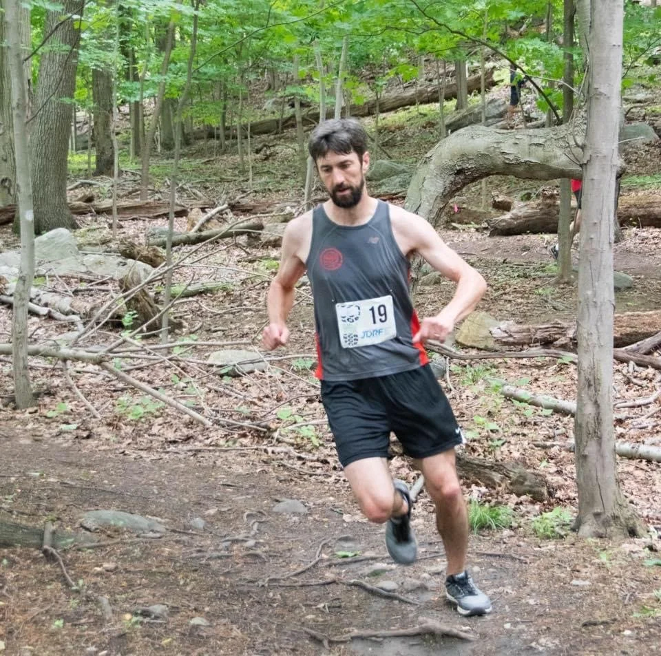A man running on a trail in a wooded forest during the pain to paine trail half marathon race, wearing a race bib with the number 19, a dark sleeveless athletic shirt, and shorts.