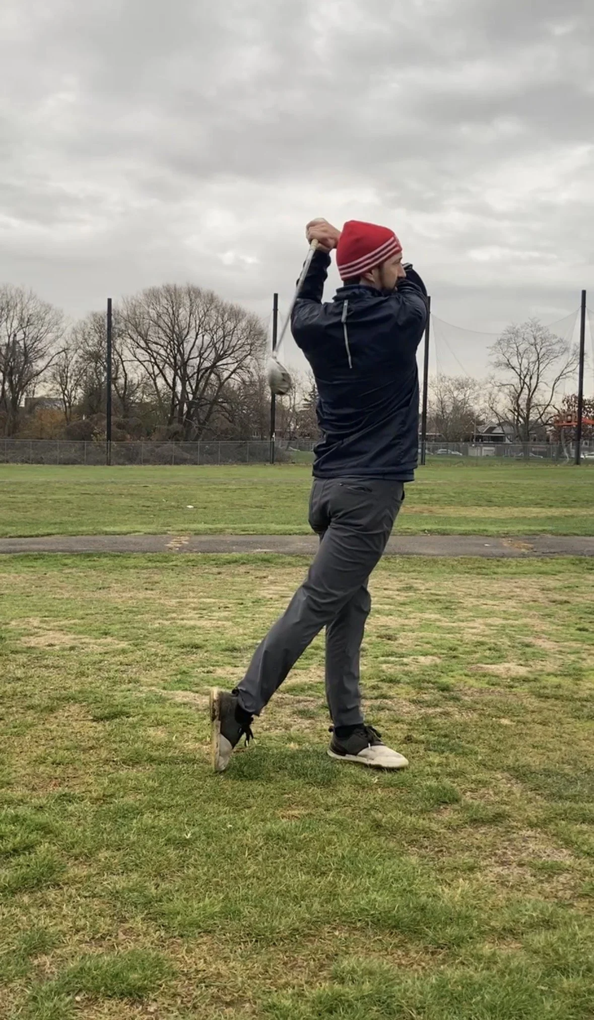 A man in a red beanie, black jacket, and gray pants is swinging a golf club on a grassy field, with leafless trees and a cloudy sky in the background.